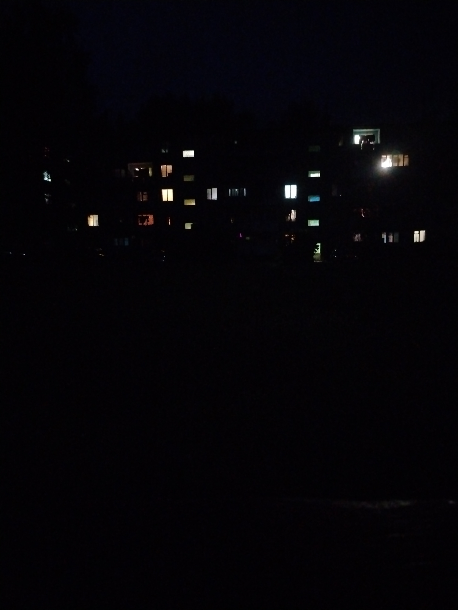 The image depicts a dark, nighttime scene of apartment buildings in Stroitel', Russia, with various windows illuminated by interior lights. The foreground is entirely black, suggesting a dark open space, possibly a field or park. The apartment buildings are silhouetted against the night sky. Several windows are visible on the facades of the buildings, with light emanating from them, indicating occupancy. The lights vary in color, with some appearing yellow or warm white, while others are a cooler white or even have a faint blue or green hue. The buildings appear to be multi-story residential structures, typical of urban or suburban environments. The overall impression is one of a quiet, residential area at night.