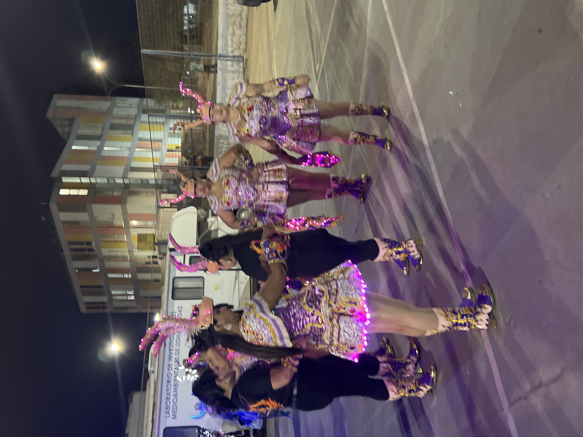 In Arica, Chile, a group of four women are standing in a paved outdoor area at night. They are dressed in elaborate, sparkly costumes with matching horned headdresses. The costumes are predominantly purple and gold, featuring intricate patterns and what appear to be LED lights around the hem of their skirts and on parts of their boots. Two of the women are wearing black tops and pants, while the other two are in matching elaborate costumes. They are positioned around a white trailer or vehicle that has text on its side, possibly indicating a laboratory or research facility. In the background, a modern building with colorful horizontal stripes is visible, along with streetlights and a fence. The lighting suggests an event or performance is taking place, with purple and pink hues illuminating the scene.