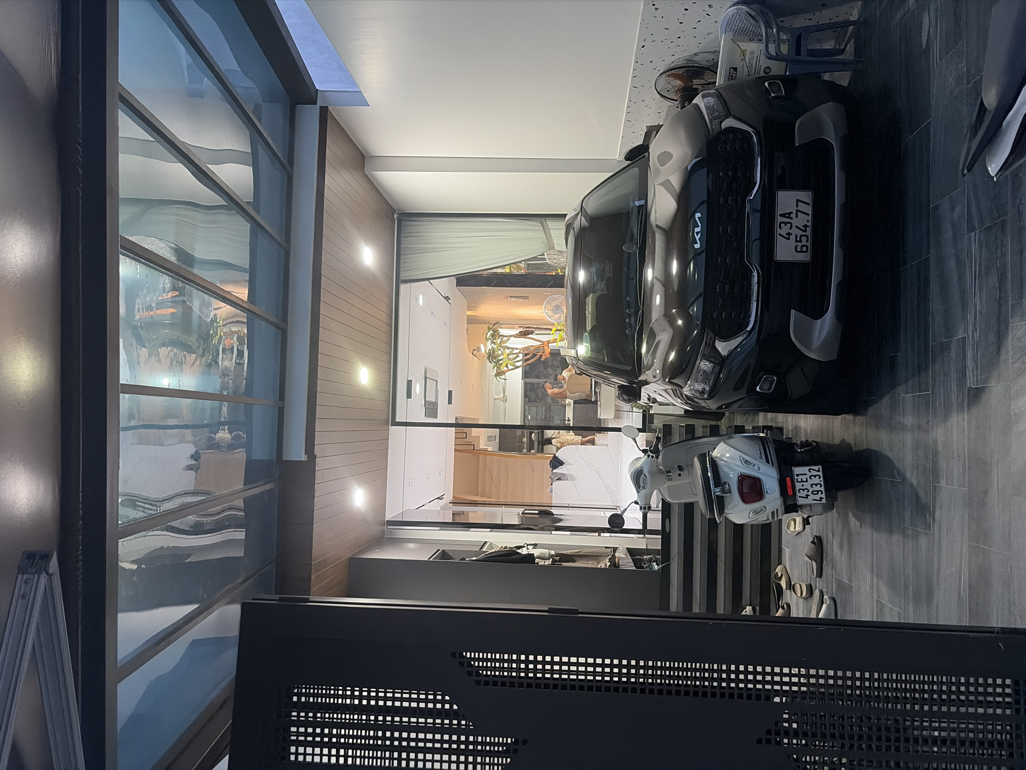 The image depicts a residential entryway in Da Nang, Vietnam, featuring a modern aesthetic. In the foreground, a black Kia Sonet is parked, its license plate reading "43A 654.77". To the left of the car, a white scooter is parked, with its license plate reading "43-E1 493.32". The scooter has several pairs of sandals scattered on the steps behind it. The entryway is covered by a transparent roof structure, allowing natural light to filter in and reflecting the surroundings. Inside the house, a staircase with wooden steps is visible, leading up to living quarters. In the background, two individuals are casually interacting within the well-lit interior, suggesting a domestic setting. The overall environment appears clean and well-maintained, consistent with urban residential architecture in Vietnam.