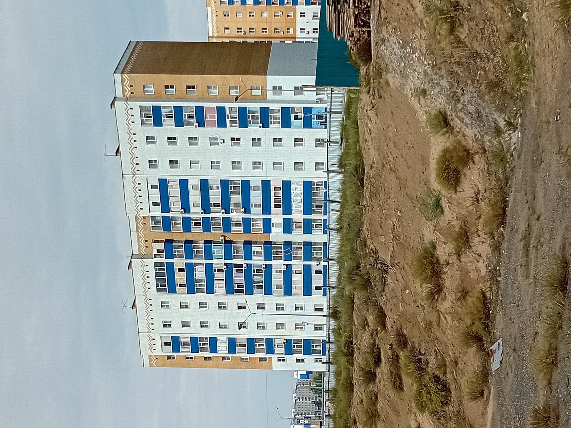 The image shows a multi-story residential building in Semey, Kazakhstan. The building is a light blue and tan color, with blue accents around the windows. It has a patterned design along the roofline. There are multiple windows visible on each floor, with some air conditioning units mounted outside. In front of the building, there is a chain-link fence and some sparse vegetation growing on a dirt embankment. In the background, other residential buildings are visible, as well as some streetlights. A pile of lumber is stacked to the right of the building, near the fence. The sky is overcast. The scene depicts an urban residential area.