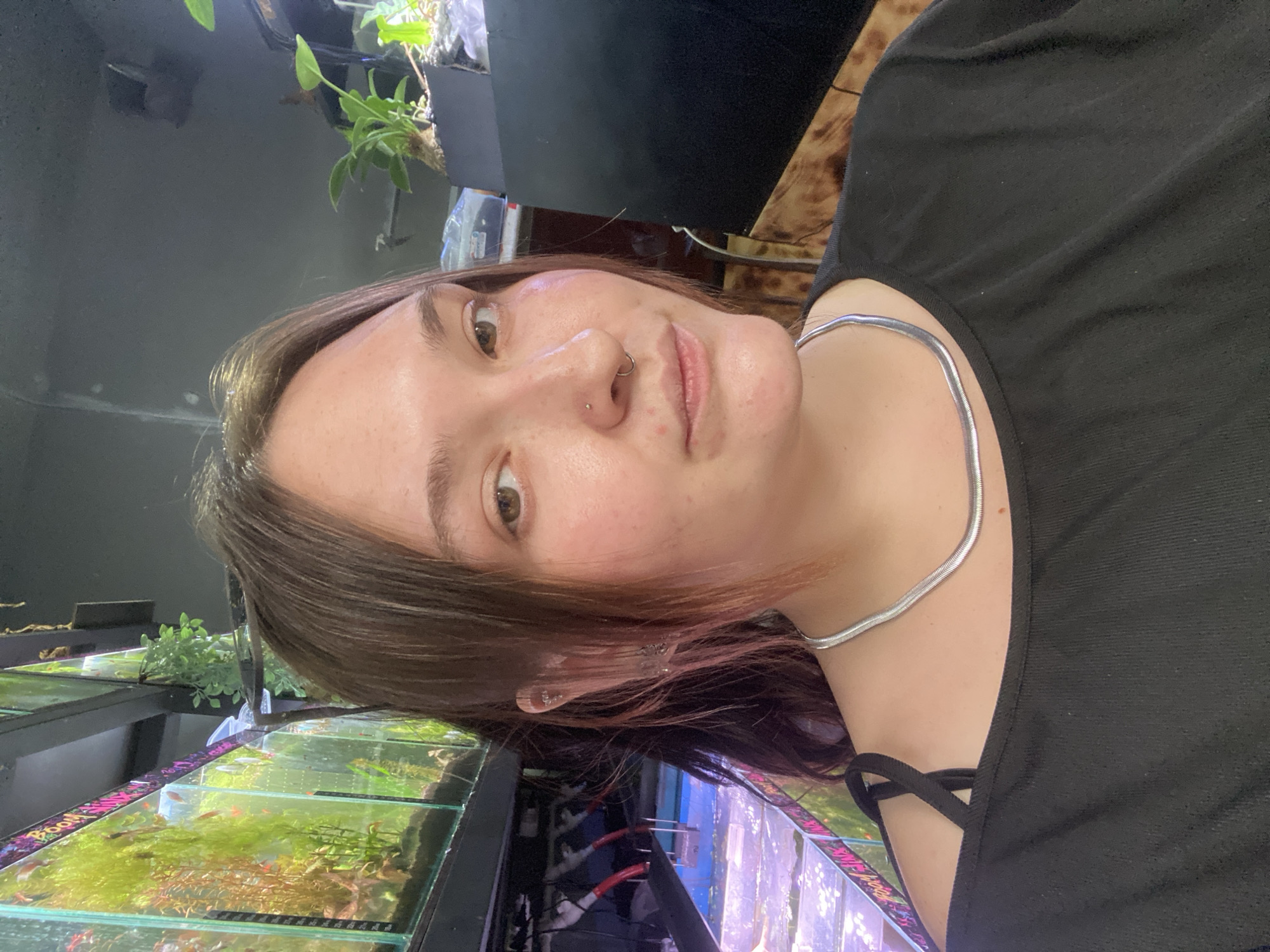 A young woman with brown hair and brown eyes is pictured from the chest up, looking towards the camera. She is wearing a black off-the-shoulder top and a silver chain necklace. She has a nose ring and piercings in her left ear. In the background, there are multiple rows of aquariums filled with water and aquatic plants. The aquariums are lit from above and appear to be in a store or public space. The overall environment is dimly lit, with the aquariums providing the primary light source. The location is not identifiable as being in Bishkek City, Kyrgyzstan, or any other specific location.