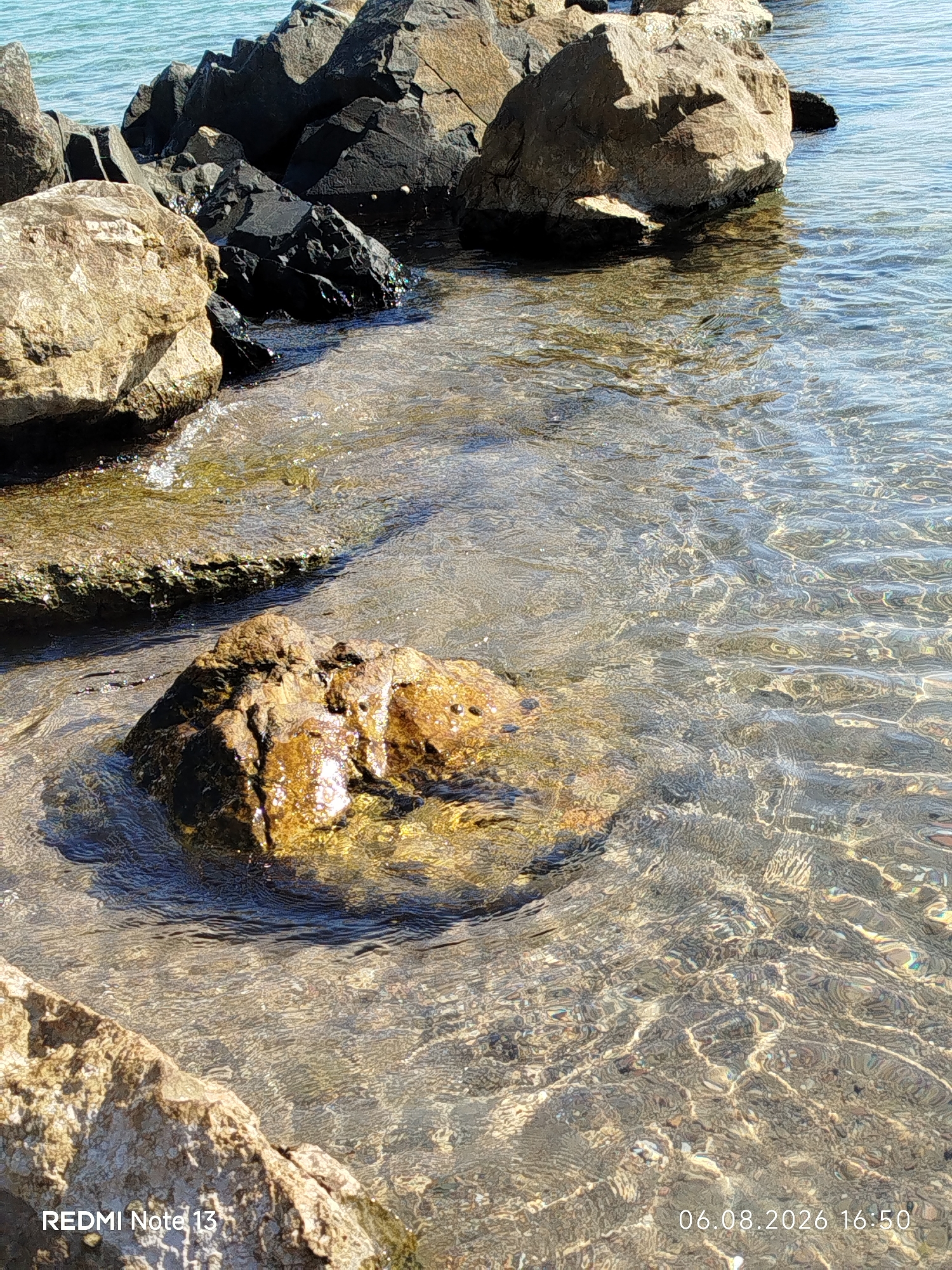 The image displays a close-up view of a rocky shoreline with clear, shallow water. Several large rocks, varying in color from light brown to dark gray and black, are partially submerged or rest on the edge of the water. The water is transparent, allowing the sandy and rocky seabed to be visible. Sunlight refracts through the water, creating shimmering patterns on the rocks and the seabed. The environment appears natural and serene, with no people or specific structures visible. The timestamp "06.08.2026 16:50" is superimposed on the lower right corner of the image, and "REDMI Note 13" is on the lower left. The scene is consistent with coastal areas in Sithonia Municipal Unit, Greece, known for its rugged coastlines and clear waters.