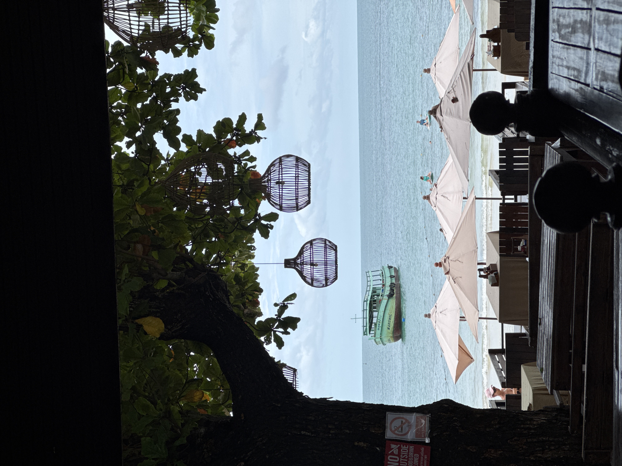 This is a full shot from a restaurant balcony looking out at the ocean in Ao Phai, Thailand. In the foreground, there are wooden tables and benches. Overhead, there are leafy trees with hanging wicker lamps. To the left, a tree trunk has two signs attached to it. Further out, a green boat with a white cross on its roof is anchored in the water. The beach is visible with several umbrellas and people on paddleboards in the ocean. The sky is overcast.