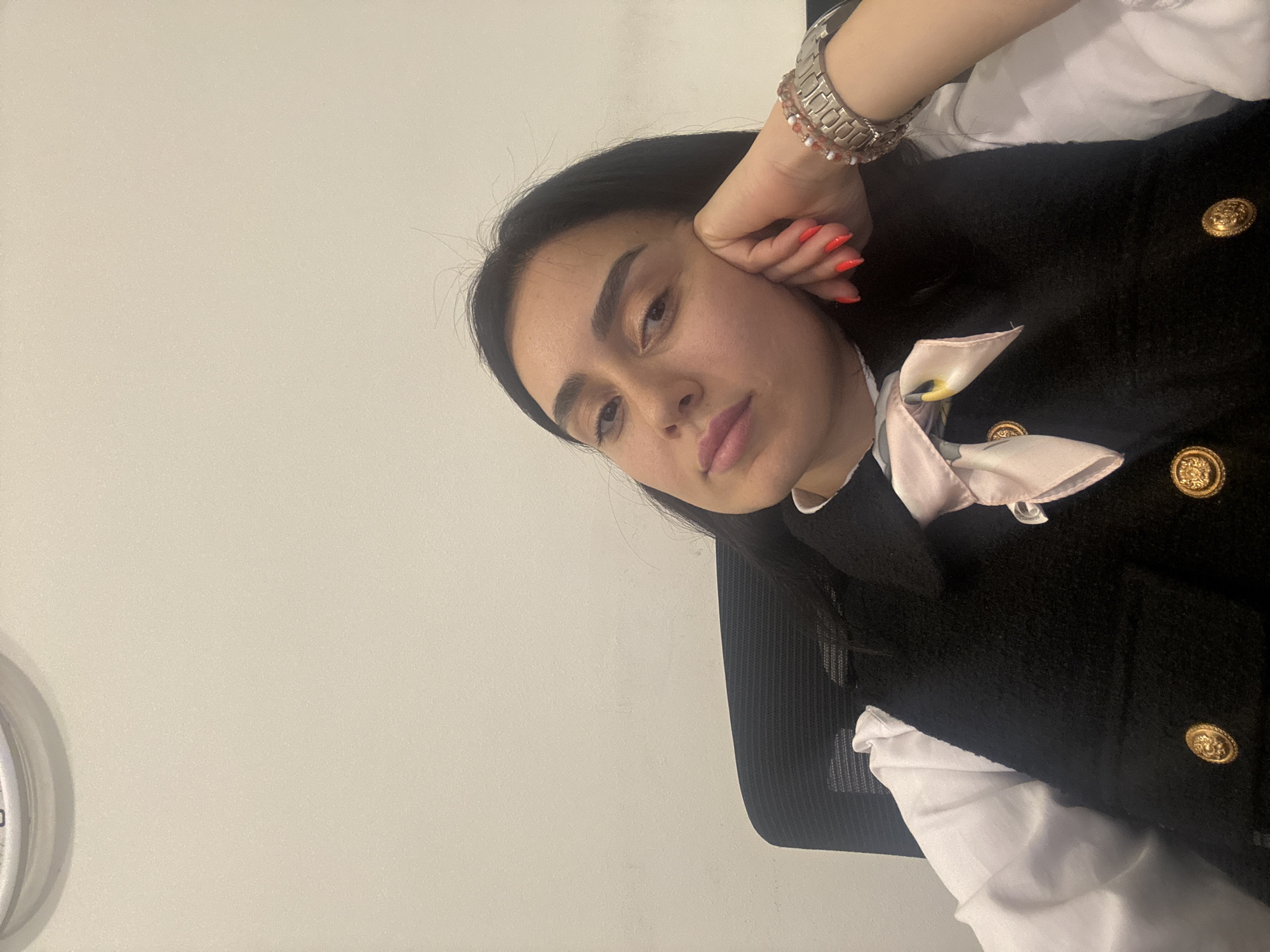 This is a portrait photograph of a young woman taken indoors, possibly in an office or professional setting in Tbilisi, Georgia. She is positioned off-center, with her head tilted, and her gaze directed towards the viewer. The woman has long, dark hair and prominent dark eyebrows, with her lips appearing to have a soft pink or nude lipstick.
She is dressed in a white collared shirt, layered under a black, textured waistcoat or jacket adorned with large, ornate gold-colored buttons. A light pink and white patterned scarf or cravat is tied around her neck. On her left wrist, she wears a silver-toned watch with a broad band and a beaded bracelet featuring red and white beads. Her fingernails are painted a bright orange.
Her left hand is raised, with fingers gently curled and resting against her cheek, contributing to a thoughtful or pensive expression. Behind her, a black office chair with a mesh back is partially visible. The background is a plain, light-colored wall, providing a clean and uncluttered setting. A small section of a metallic, circular object, potentially a clock, is visible on the far left edge of the frame. The lighting is even, suggesting an indoor environment with controlled or diffused light. No text is visible in the image.