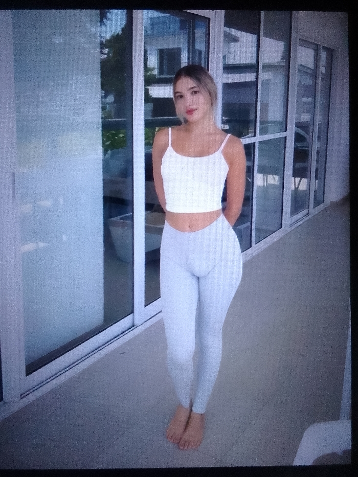 The scene depicts a young adult female standing barefoot on a light-colored paved outdoor surface, consistent with a balcony or patio, in the city of Unknown, Unknown. She is positioned centrally, facing the camera. The subject has light brown hair and a fair complexion. She wears a white ribbed spaghetti-strap crop top, exposing her midriff, where a navel piercing is visible. Her lower body is clad in light gray, form-fitting leggings. Her hands are clasped behind her back. The background consists of floor-to-ceiling glass sliding doors with white frames, reflecting elements of modern architecture and green foliage, indicating a contemporary residential or hotel setting. The lighting suggests daytime.