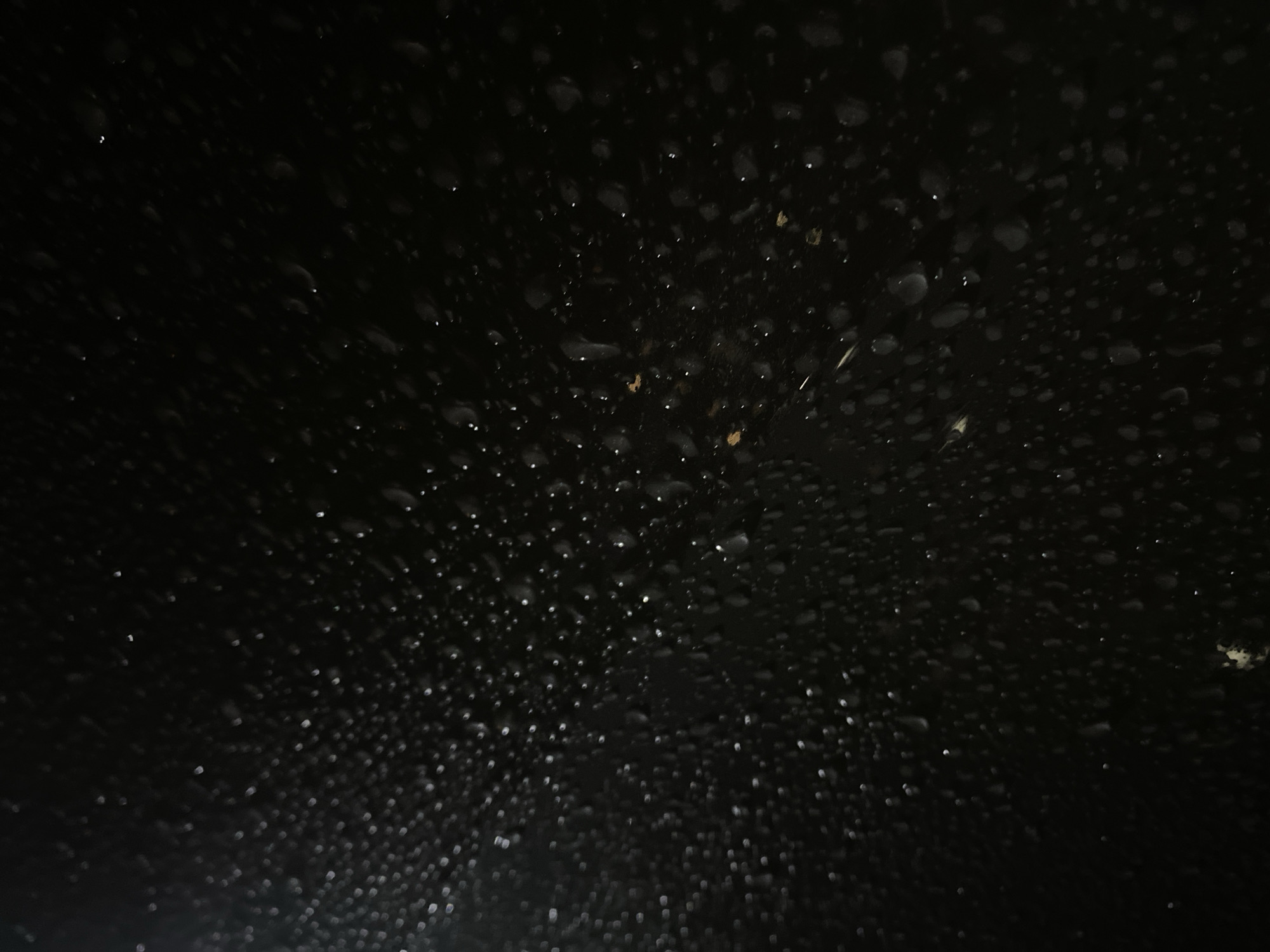 The image is a close-up, dark, and somewhat blurry photograph of water droplets on a dark, textured surface. The droplets vary in size, with some appearing as small, distinct dots and others as larger, more amorphous shapes. The lighting is dim, with some areas appearing brighter due to reflections off the water. There are a few small, light-colored specks that could be dust or debris. The background is uniformly dark, obscuring any details of the surrounding environment. There are no people, animals, or recognizable objects visible. The image was taken in Balashikha, Russia.