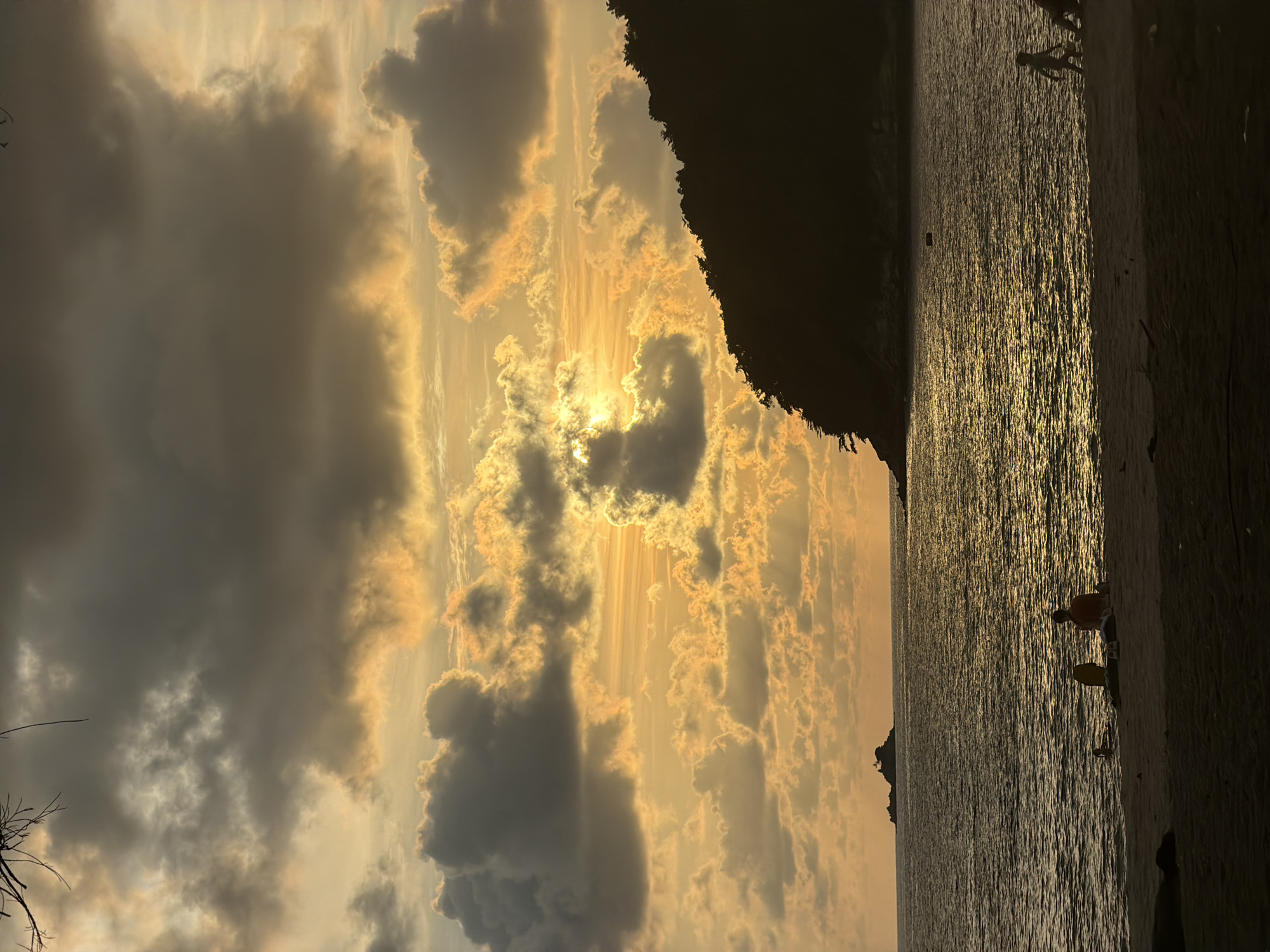 The image shows a golden sunset over the ocean, likely in Choeng Thale, Thailand. Heavy clouds obscure the sky, but the sun shines brightly through, creating a dramatic light display. To the right, a dark landmass with dense foliage juts into the scene. The ocean reflects the golden light of the sun, creating a shimmering effect. In the lower right corner, two figures are visible on the beach. One figure is crouched down near the water's edge, and another stands behind them. A third figure is further out in the water. The overall tone of the image is serene, showcasing the natural beauty of the location.