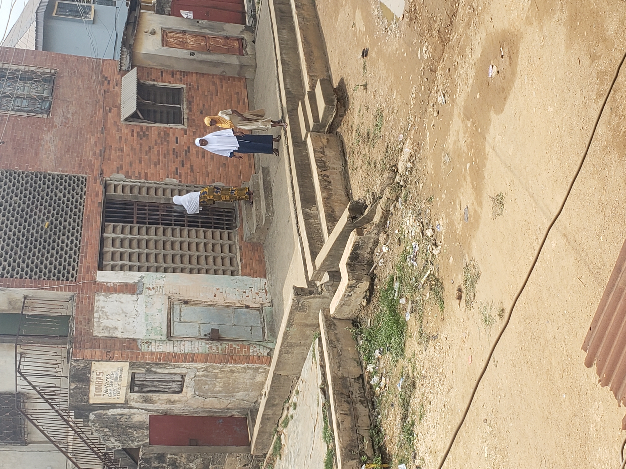 The image captures an outdoor scene in what appears to be a residential area in Alalubosa, Nigeria, under bright, clear daylight. The ground, predominantly sandy dirt with sparse patches of green vegetation and scattered litter, occupies the right side of the frame. A dark cable or wire runs across this unpaved ground.
A series of worn concrete steps and raised pathways traverse the middle ground, leading up towards the left. On the left side, multi-story buildings constructed from red brick and concrete dominate, exhibiting signs of age with some faded paint, exposed brickwork, and general wear. The buildings feature various window designs, including some with metal security grates, wooden shutters, and sections of patterned concrete block walls. A rusty metal staircase or railing is visible on the lower left, attached to one of the buildings.
Three individuals, all appearing to be women wearing traditional attire with head coverings, are present. One woman, in a white head covering and dark blue attire, is positioned on the concrete steps, facing right. Another woman, wearing a yellow head covering and a light-colored dress, stands slightly behind her near the steps, also facing right. A third woman, in a white head covering and a patterned skirt, is partially visible on the far left, near the base of one of the buildings. They appear to be walking or standing along the pathway.
On a building wall on the lower left, a white rectangular sign with black text is visible. The legible text includes "TONBAS Ventures," "GSS SERVICES," and an address containing "ZAZAA R.D. OLUWA NAJA." A piece of corrugated metal, possibly part of a fence or roof, is seen on the far right.