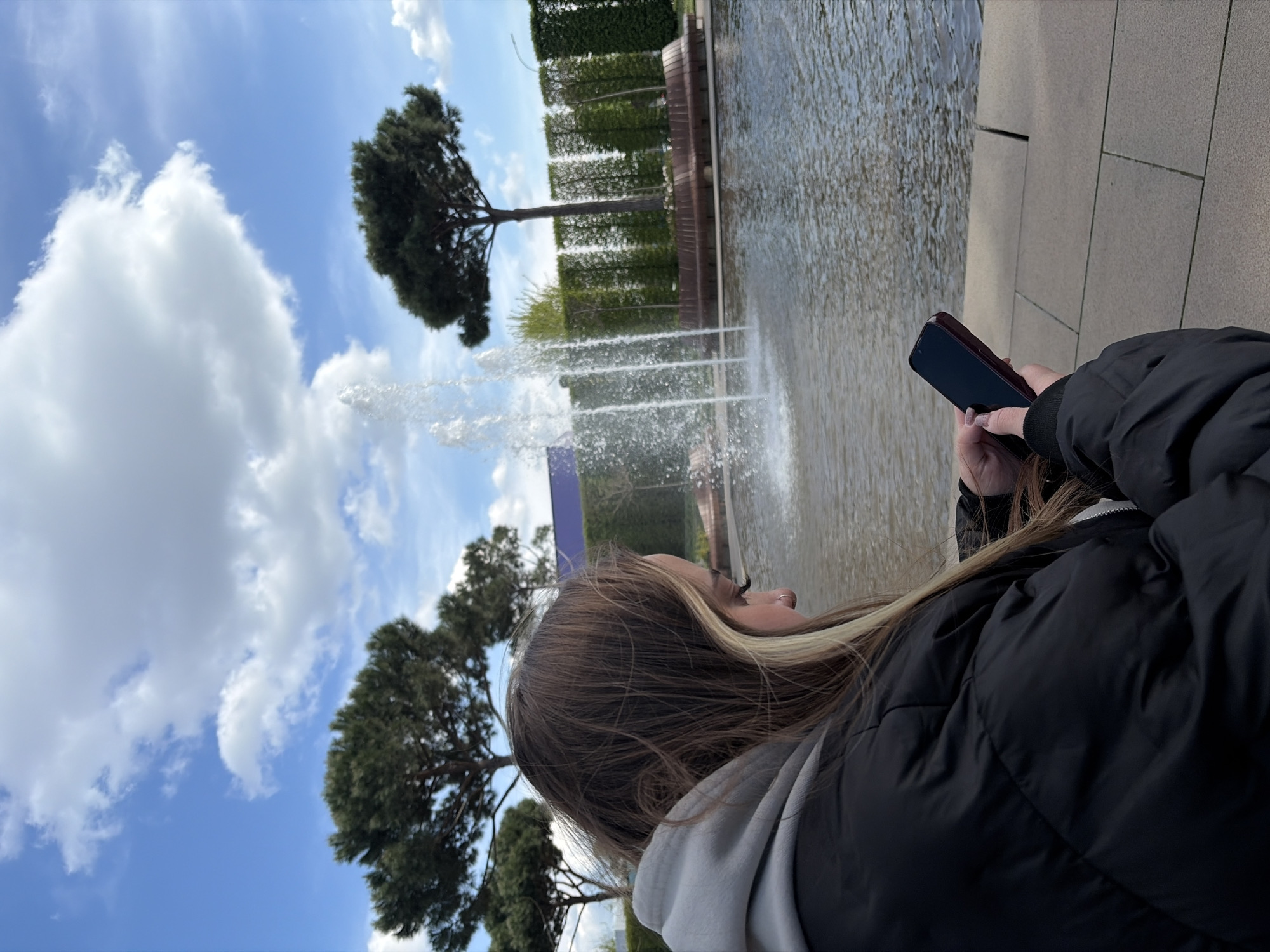 The image presents an outdoor scene, likely within a public park or garden in Krasnodar, Russia, captured on a bright, partly cloudy day. The shot is taken from behind and to the right of a person, who is observing a water feature.
The main subject is a young person with long brown hair featuring blonde highlights, seen in profile. They are dressed in a black puffer jacket worn over a light gray hooded sweatshirt. Their right hand holds a dark-colored smartphone, with manicured nails visible. The person's gaze is directed forward, seemingly towards the water fountain.
In the mid-ground, a series of powerful water jets spray upward from a large body of water, possibly a pond or a wide fountain pool. To the right of this water feature, a walkway paved with light gray rectangular stones is visible, indicating the ground where the person is standing.
The background is rich with greenery: several large, dark green trees with dense, rounded canopies are prominent on the left side, with another tree further back. Behind the water and extending along its edge, tall, neatly trimmed green hedges form a structured wall, with subtle wooden elements interspersed. The sky above is a clear blue, adorned with scattered white cumulus clouds, suggesting pleasant daytime weather. No visible text is present in the image.