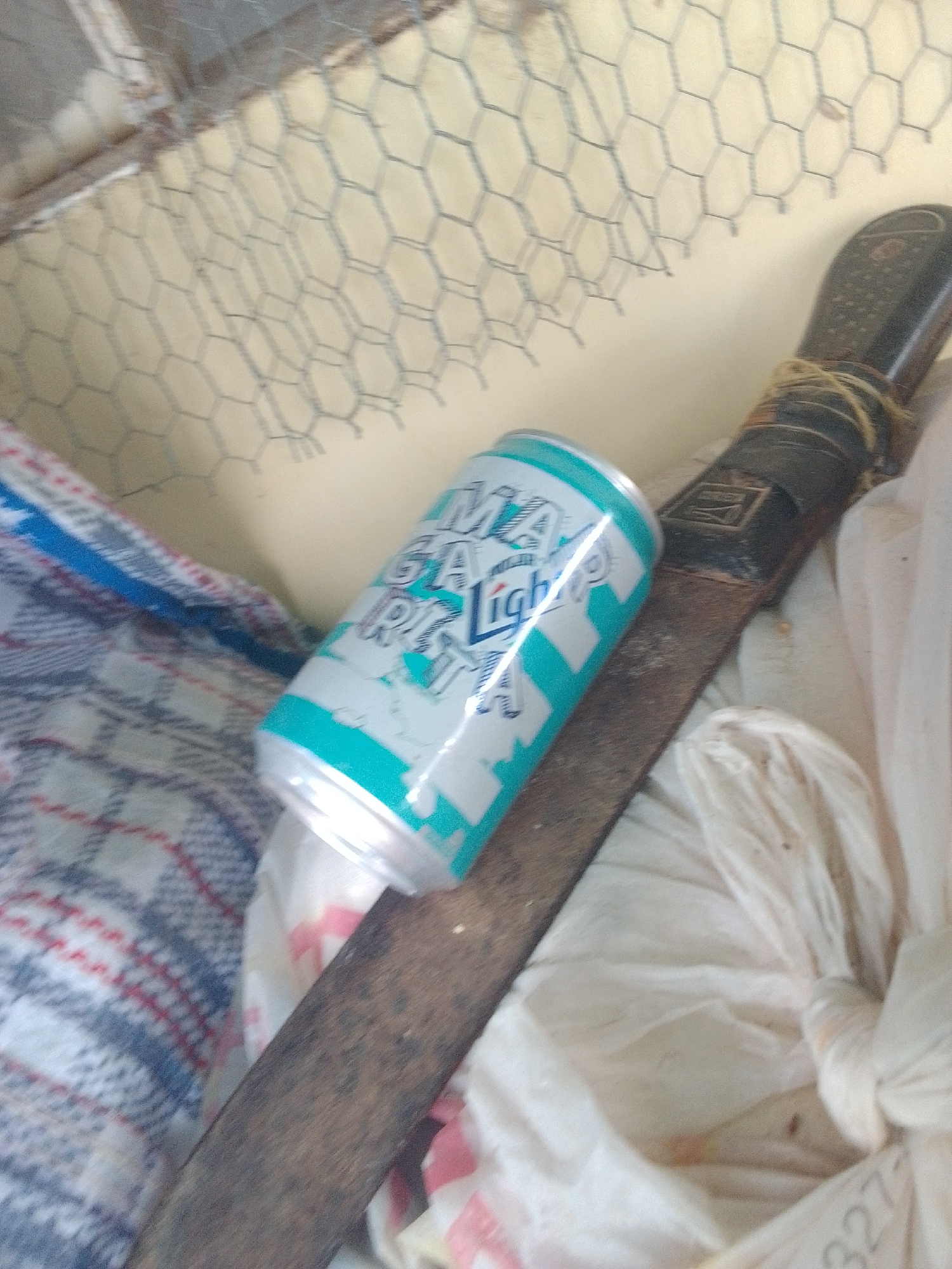 The image shows a close-up view of several objects, likely in a dimly lit, indoor or sheltered setting. In the foreground, a can with a turquoise and white design is partially visible. Text on the can, though partially obscured and blurry, appears to read "GA Light." To the right of the can, a weathered machete or large knife is laid horizontally. Its handle is dark, with a string tied around the top portion. The blade shows signs of rust and use. Behind the can and machete, a textured surface, possibly chicken wire or a similar mesh material, is visible against a pale yellow background. To the left of the can, a folded piece of fabric with a checkered pattern of blue, white, red, and gray is present. Further to the right, a white plastic bag, possibly filled with something, is loosely tied. The overall impression is one of clutter or storage, with no clear indication of a specific activity or event. The lighting is subdued, and there are no discernible environmental cues to suggest time of day or weather. The location context is La Tagua, Venezuela.