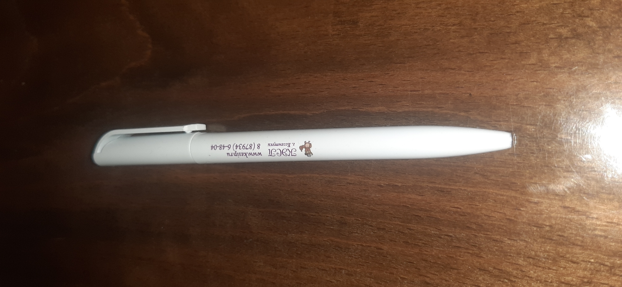 A white pen lies on a wooden surface. The pen has text printed on it in a dark reddish-brown color, along with a small illustration of what appears to be a donkey or mule. The text on the pen reads "КСП www.kesip.ru г. Ессентуки 8(87934) 6-48-04". This indicates that the pen is associated with an organization located in Yessentukskaya, Russia, and provides a website and phone number. The wooden surface appears to be a table or desk, with visible grain and some sheen reflecting light. The lighting in the image is dim, casting shadows on the surface.
