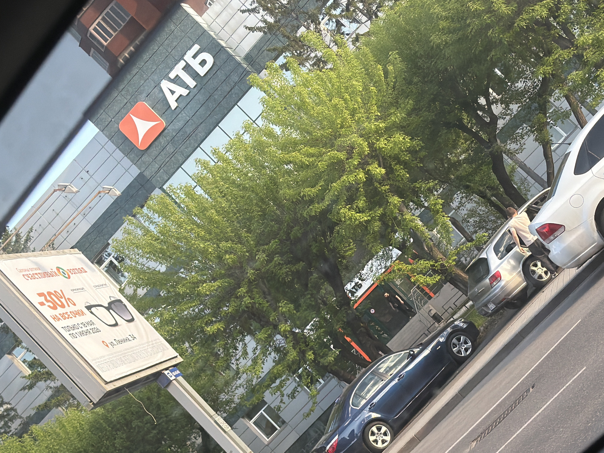 The image shows a street scene in Krasnoyarsk, Russia, featuring the "ATB" bank building in the background. A large advertising sign for "Счастливый взгляд" (Happy View) is prominently displayed, promoting a 30% discount on all glasses from May 18 to June 1, 2026, at 34 Lenin Street. Several cars are parked along the street, including a blue sedan and a silver SUV. Trees provide partial coverage of the building and street. A person is visible near one of the parked cars. The photo appears to have been taken from a car.