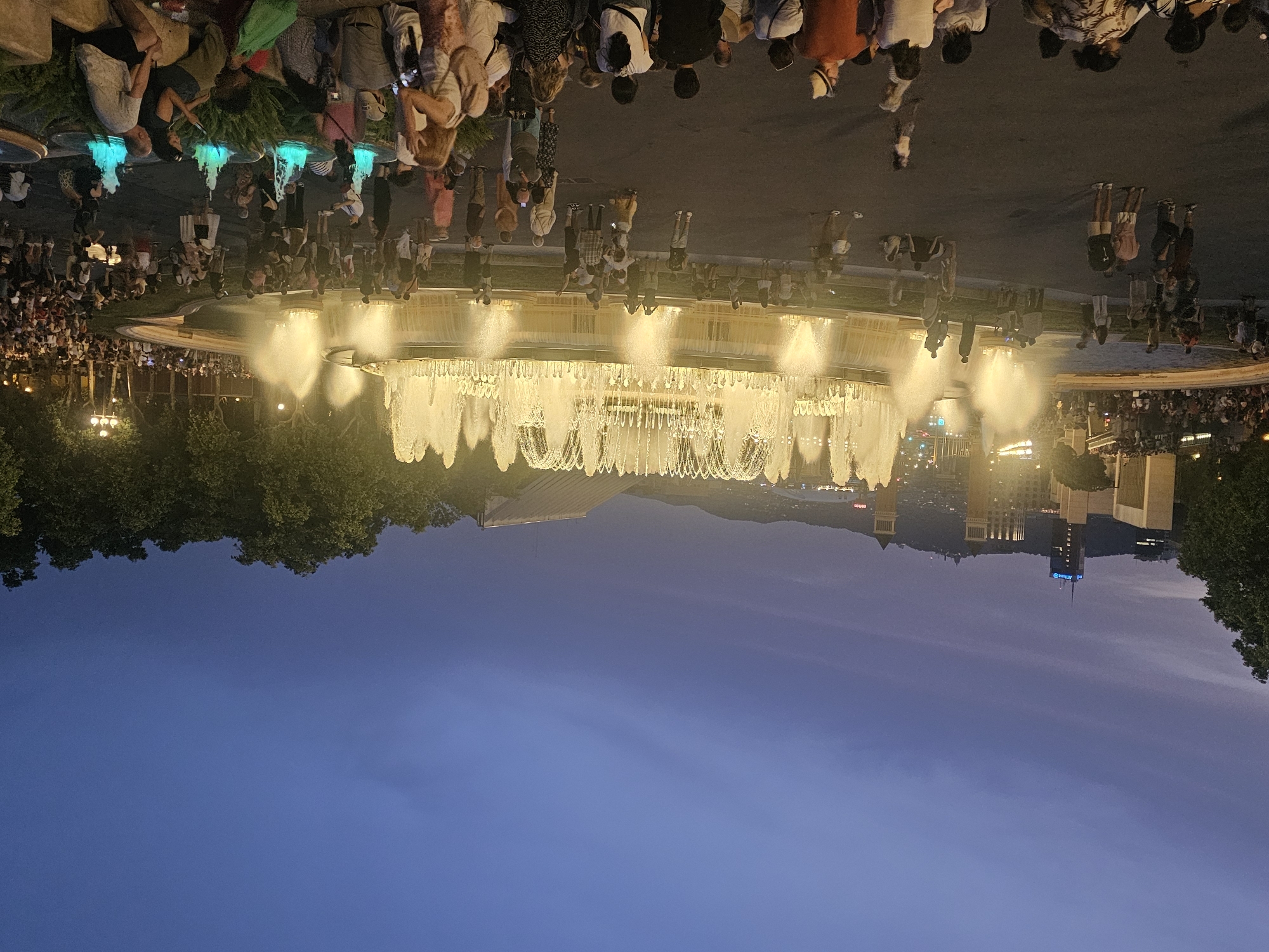 The image shows a wide shot of the Magic Fountain of Montjuïc in Barcelona, Spain, at dusk. The fountain is illuminated with bright, golden light, with numerous jets of water spraying upwards and outwards. A large crowd of people are gathered around the fountain, watching the display. Some people are standing, while others are sitting on the ground. The sky above is a deep blue, with some clouds. In the background, the cityscape of Barcelona can be seen, with buildings and lights. The overall atmosphere is one of a public spectacle and gathering.