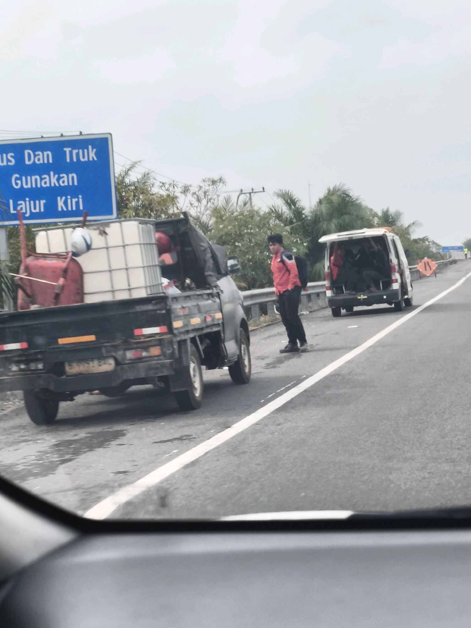A person in a red shirt and black pants stands on the side of a road, facing a white van. The van's rear doors are open, revealing several people inside. In front of the van and slightly to the left is a black pickup truck carrying a large white container and a red wheelbarrow. A blue sign with white text is visible to the left of the pickup truck, indicating "Bus Dan Truk Gunakan Lajur Kiri" (Buses and Trucks Use Left Lane). The scene takes place on a multi-lane road with a white line dividing the lanes. The environment appears to be outdoors with trees and a cloudy sky. The location is identified as Pemulutan Ulu, Indonesia.