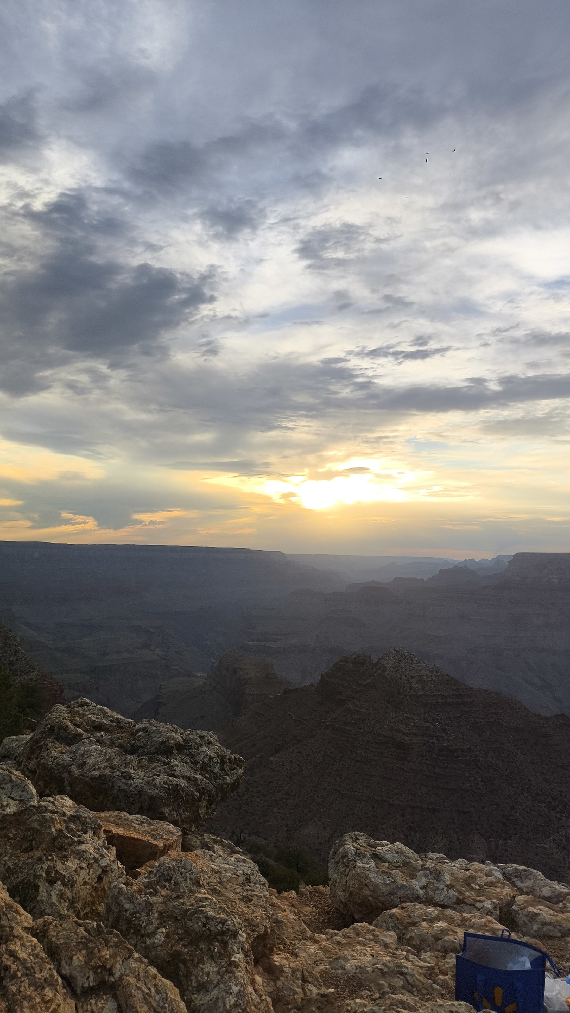 A vertical, full-shot image captures the Grand Canyon at dusk. The sky is dominated by dramatic clouds, with a bright band of sun peeking through the center, casting rays down onto the canyon. The rugged terrain of the canyon, layered in shades of gray and brown, stretches into the distance. In the foreground, large, textured rocks and boulders fill the bottom of the frame, some with hints of orange and yellow hues. To the far right, a blue reusable shopping bag rests on the rocks. Several birds are visible as tiny specks against the cloudy sky. The scene appears to be from a viewpoint overlooking the canyon, likely on the South Rim near Grand Canyon Village, Arizona, United States.