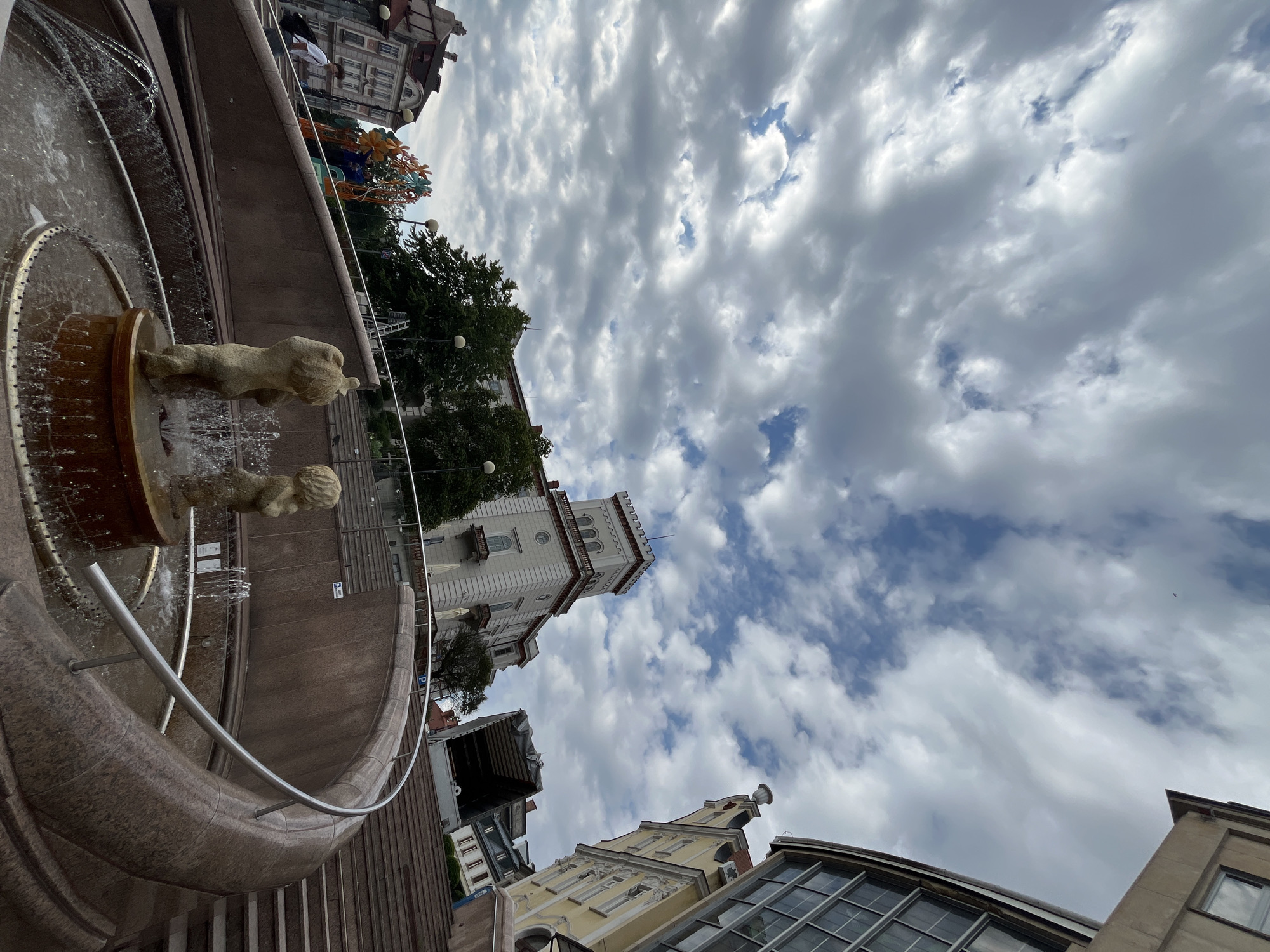 This low-angle, wide shot captures a fountain in the foreground with two cherubic statues spouting water. A curved granite wall surrounds the fountain, leading up to a wide staircase on the right. In the midground, a large, light-colored building with a tower stands prominently, flanked by trees and other architectural structures. The sky above is filled with dramatic, textured clouds. The scene appears to be an urban public space, possibly a square or park, with a mix of historical and modern architecture. The fountain's design features a rustic base and spouting cherubs, while the surrounding architecture suggests a European city.