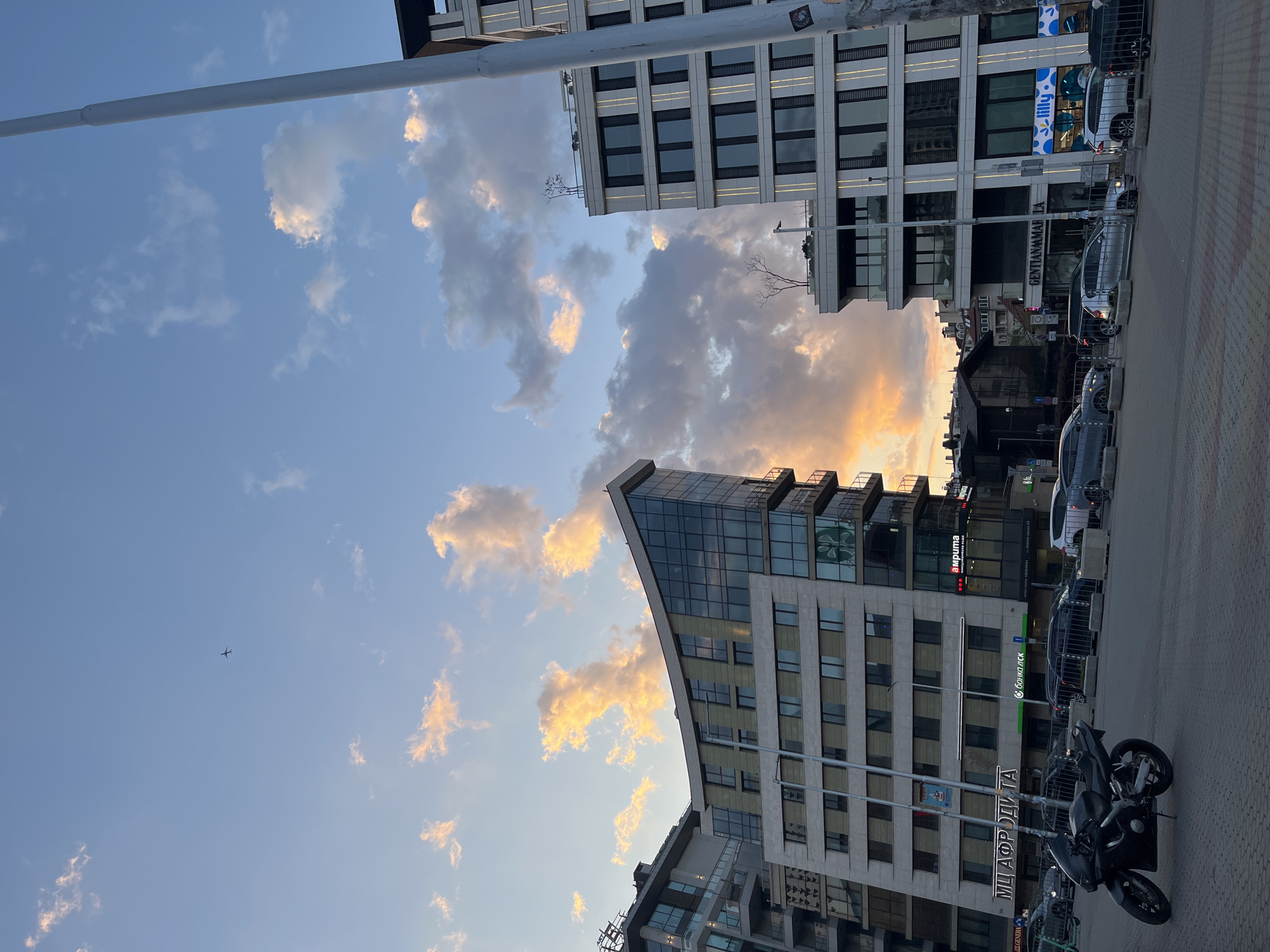 The image shows a street scene in Sofia, Bulgaria, with buildings and a partially cloudy sky. The buildings have a modern architectural style, with glass and concrete facades. Several cars are parked along the street, and a motorcycle is in the foreground. Signage in Bulgarian is visible on the buildings. The sky is blue with white and orange-tinted clouds, suggesting either sunrise or sunset. A small airplane is visible in the distance.