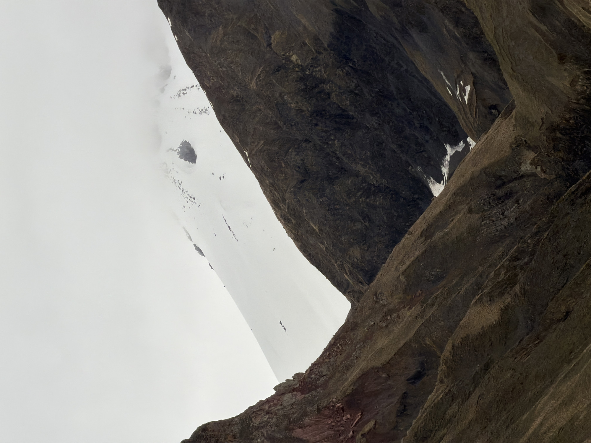 The image shows a low-angle view of a mountainous landscape. Prominent rock formations dominate the right side, with visible striations and textures. The left side displays a snow-covered or cloudy area. Small patches of snow are also visible on the rock faces.