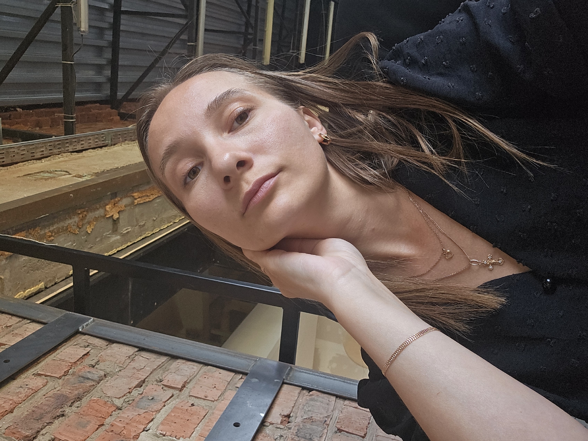 The photograph shows a young woman indoors in Chaikovsky, Russia. She has light brown hair, fair skin, and is wearing gold hoop earrings. She is wearing a black blouse or dress. A delicate gold necklace with a cross pendant is visible around her neck, along with a bracelet of the same metal on her wrist. She is resting her head on her hand. The background includes brickwork and steel beams, giving the impression of an industrial or loft-style space. There is what appears to be an interior view of a building under construction, seen behind and below the brickwork, with what appears to be building materials behind a metal grate.