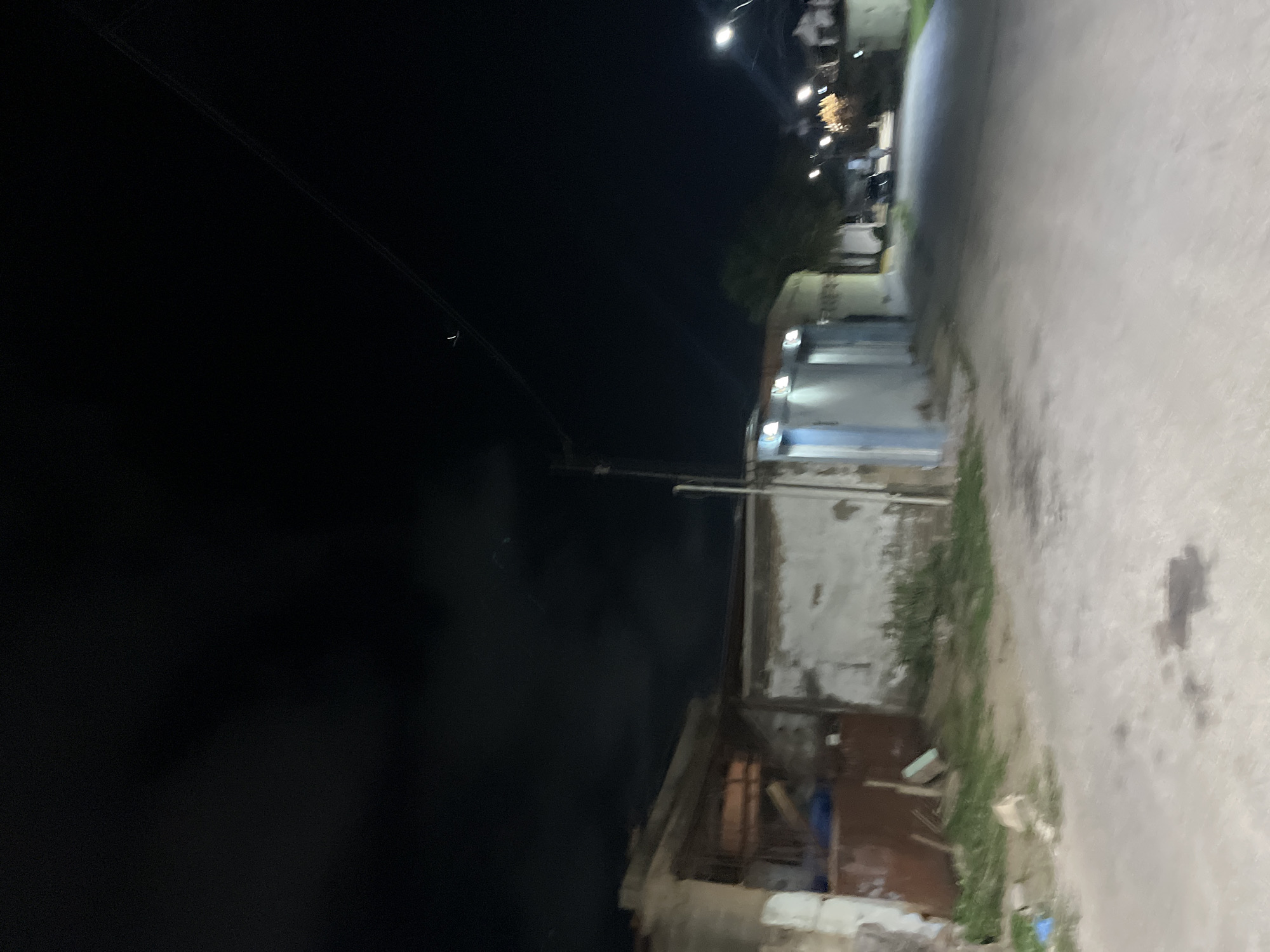 The image depicts a dimly lit street scene in Santa Ana, Venezuela, at night. In the foreground, a paved road shows some dark stains, possibly oil or water. To the left, a dilapidated structure with white and light grey walls is visible, featuring a large brown metal door or gate and an opening that reveals wooden beams inside. Some green vegetation grows along the base of this building. Further down the street, on the right, streetlights illuminate a residential area with houses and more vegetation. The sky is dark, with a few faint clouds, and power lines are visible stretching across the frame. The overall impression is one of a quiet, somewhat run-down urban environment after dark.