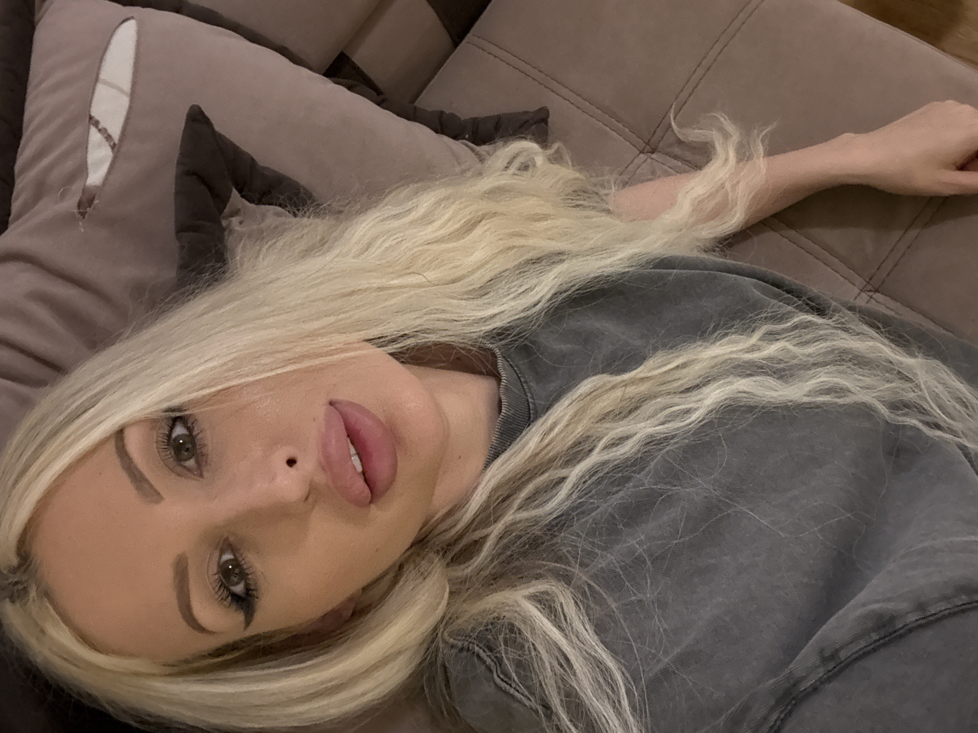 A close-up, high-angle, indoor selfie shows a woman with long, wavy blonde hair lying on a tufted brown couch. She is wearing a grey t-shirt. Her makeup includes winged eyeliner and full lips. The couch cushion behind her has a ripped section. The lighting is soft and appears to be artificial.