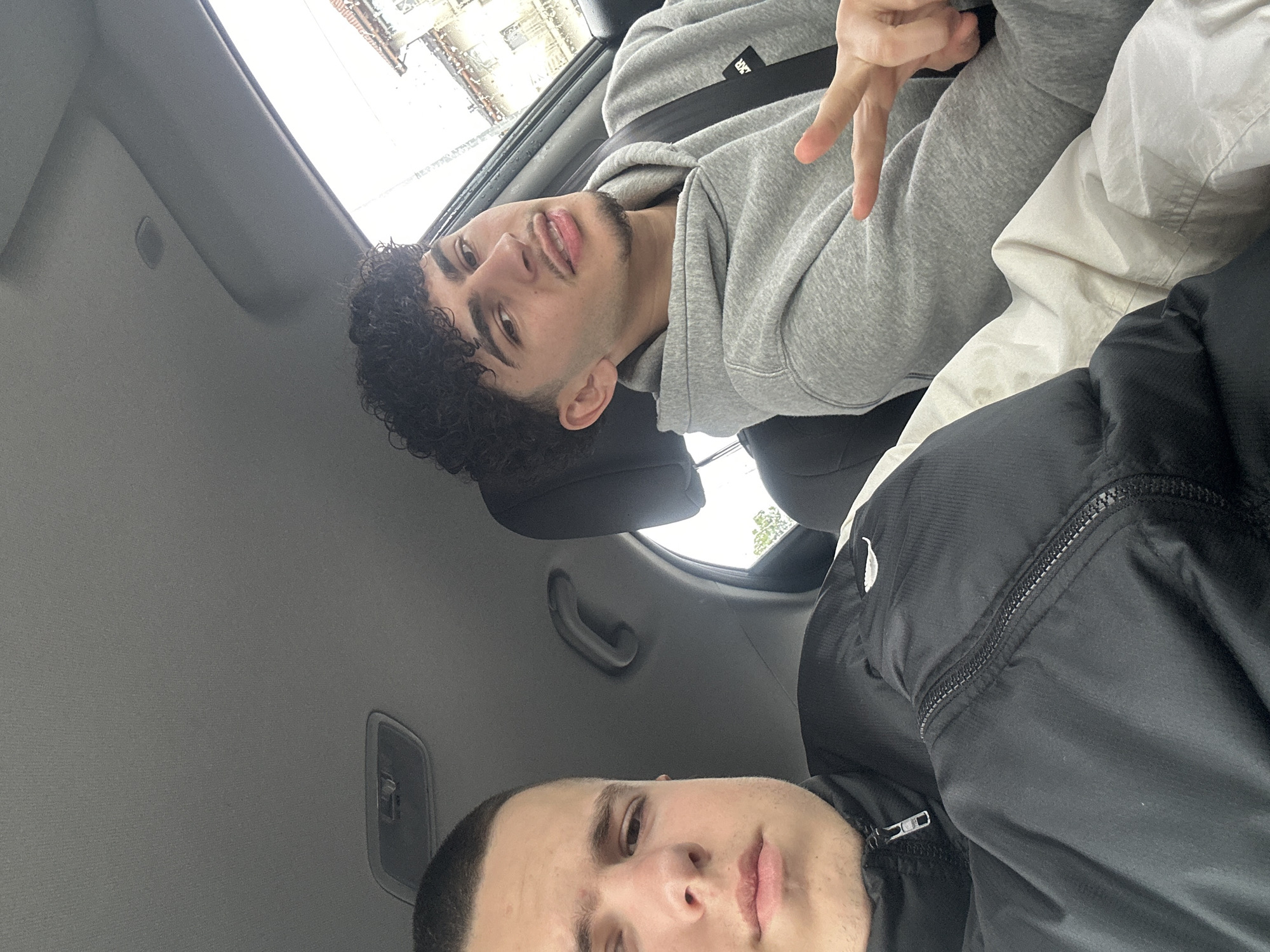 Two young men are in the interior of a car, the location of which is in Piacenza, Italy, implied but not directly visible. The camera angle is low, facing upwards towards the individuals. One man has dark, curly hair and is wearing a gray hooded sweatshirt with a visible strap across his chest. He is making a peace sign with his fingers. The other man has short, light-colored hair and is wearing a black jacket. The car's interior is visible, including the roof, a handle, and what appears to be a sunroof or window with some light and blurry shapes suggesting an urban environment outside.