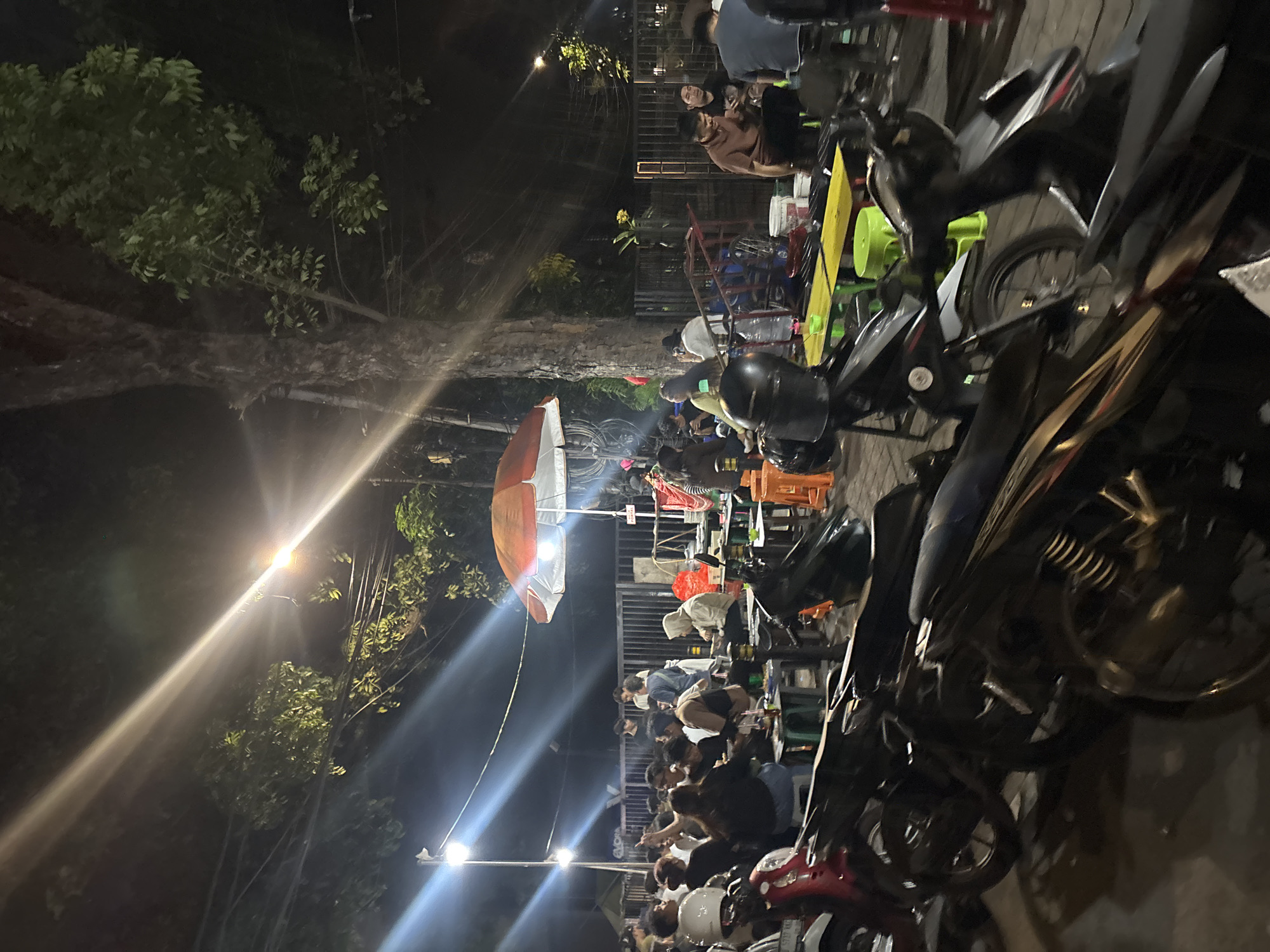 This image captures a street scene in South Jakarta, Indonesia at night. The scene is illuminated by streetlights. A group of people are gathered, seated at low tables and stools, suggesting an informal dining or socializing arrangement. An open umbrella stands amidst the tables. Several parked motorcycles are prominently positioned in the foreground, cluttering the view. Vegetation, potentially trees, looms on the left side of the frame. The composition is crowded, with an emphasis on the urban atmosphere.