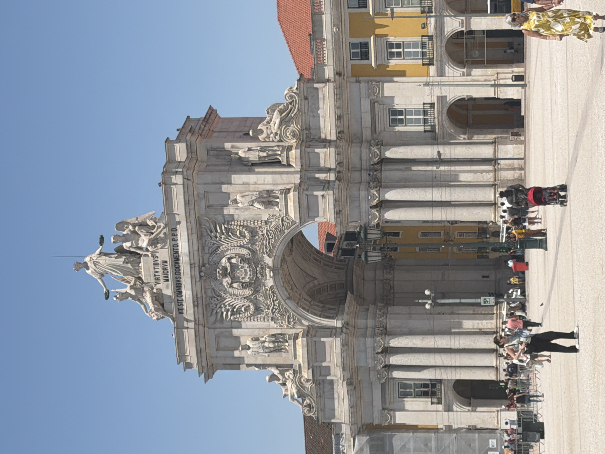 The image captures a full frontal view of the Rua Augusta Arch in Lisbon, Portugal, a majestic triumphal arch constructed from white stone. The arch stands prominently against a clear blue sky, with the sun casting shadows, indicating it is likely midday. The arch is adorned with intricate sculptures and relief carvings, with a statue of Glory at its apex, flanked by other allegorical figures. Below the apex, the inscription "VIRTUTIBVS MAIORVM" and "VT SIT. OMNIBVS. DOCVMENTO. P.P.D." are visible.
The arch opens to reveal a street with buildings visible beyond. To the right of the arch, a vibrant yellow building with orange-tiled roofs and arched windows is visible. To the left, a scaffolding suggests ongoing construction or renovation.
In the foreground, a public square is bustling with activity. Several people are walking around, with some individuals clearly in focus. A woman in a striped top and black pants stands in the center of the frame, looking away from the camera. To her left, a man pushes a stroller with a child. Further to the right, a woman in a yellow floral dress walks with a person whose hand is not visible. In the background, there are more pedestrians, lampposts, and street furniture. The ground of the square is paved with light-colored stones. The overall scene conveys a sense of a lively urban environment on a sunny day.
