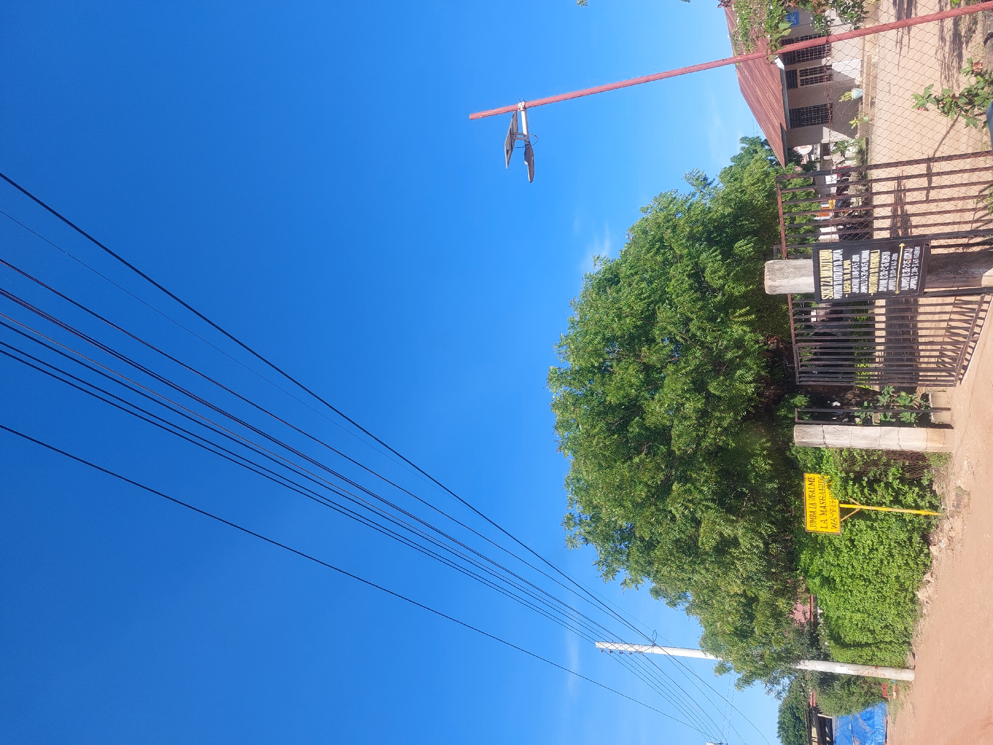 The image shows a street view in what appears to be a residential area. The sky is clear blue and visible between numerous power lines. A green tree is adjacent to a chain-link fence. Behind the fence, a building with a red roof is visible. A yellow sign displays text written in Swahili, reading "Jumba la Ufalme la Mashahidi wa Yehova." The image suggests this is near the Kingdom Hall of Jehovah's Witnesses. Given the language on the sign, the location is likely in Dar es Salaam, Tanzania.