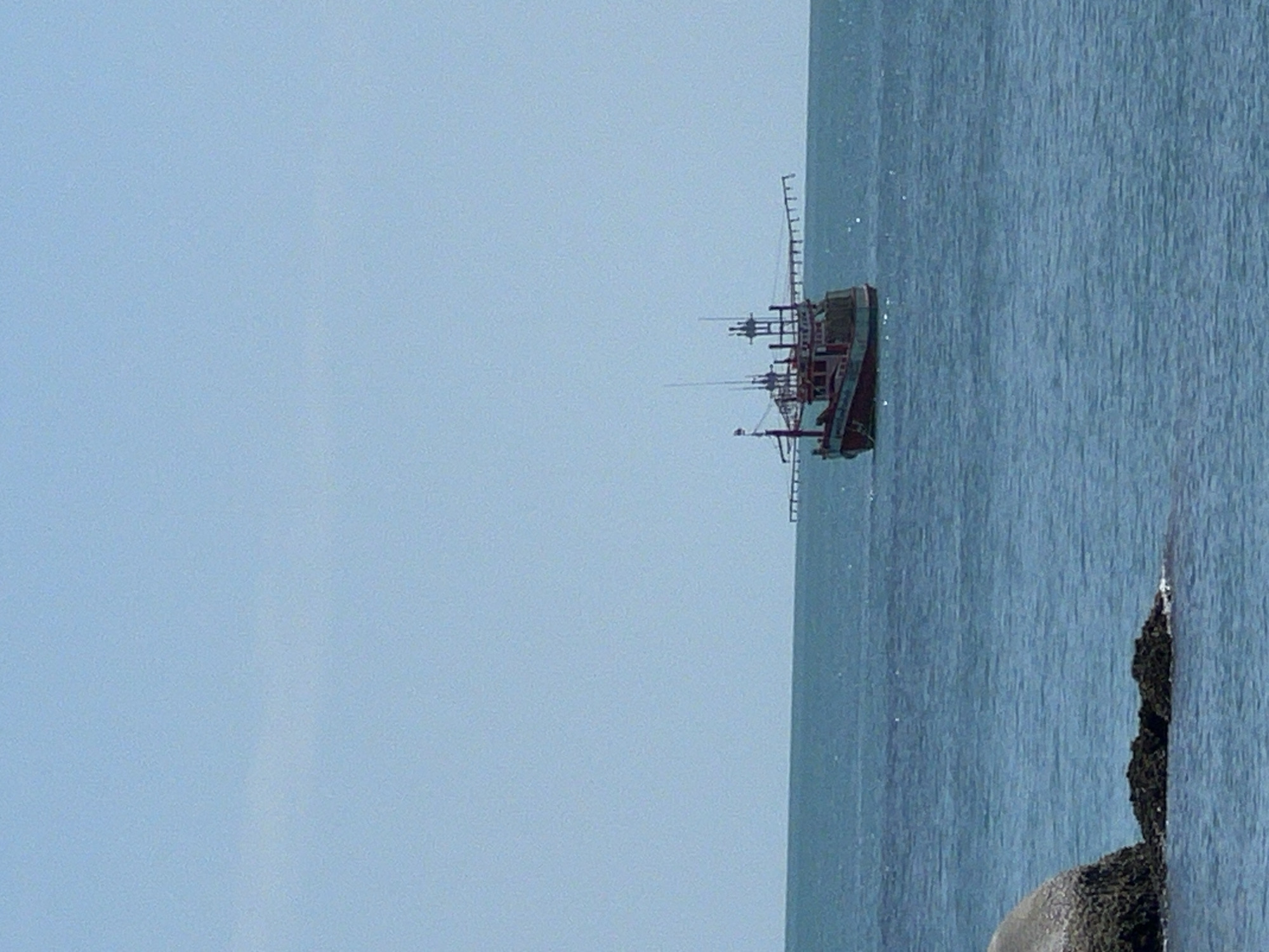A red and green fishing boat is anchored in the blue ocean under a clear blue sky. The boat has a tall structure in the center with several poles extending outwards. The horizon is visible in the distance. In the foreground, there are two large rocks partially submerged in the water. The scene appears to be in the waters off Ko Samui District, Thailand, given the distinctive style of the fishing boat and the clear, tropical waters.