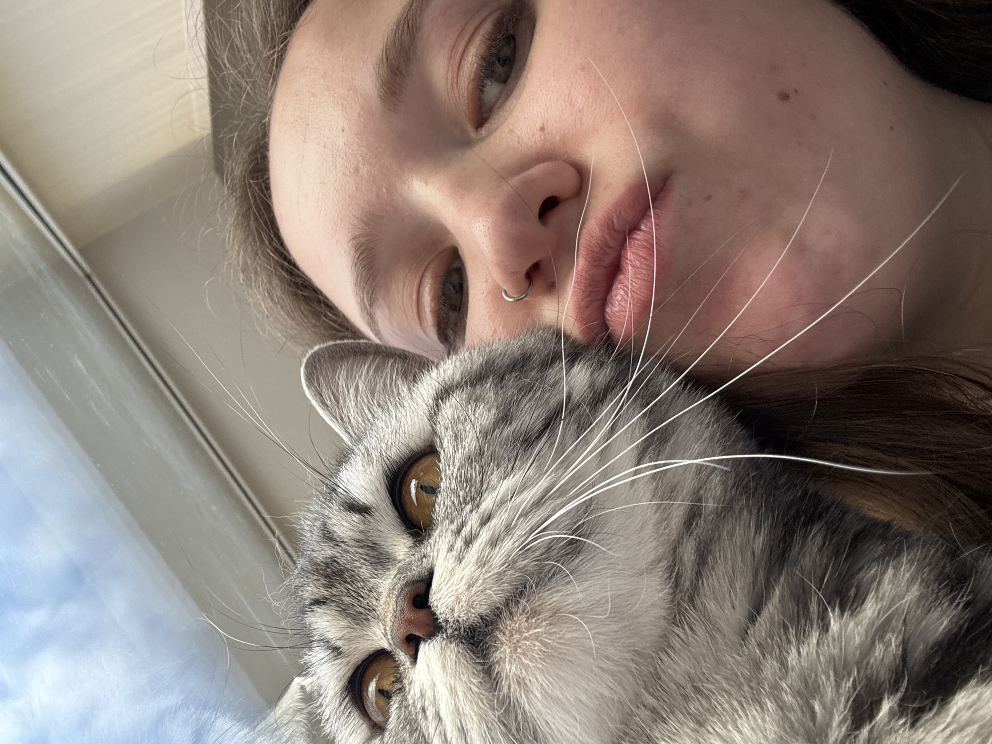 A young woman with brown hair and a nose ring is posing for a close-up selfie with a gray tabby cat. The woman is looking down at the camera, and her face is partially obscured by the cat. The cat is looking up at the camera with its amber eyes. The background is blurred. There is no evidence to suggest this image was taken in Izhevsk, Russia.