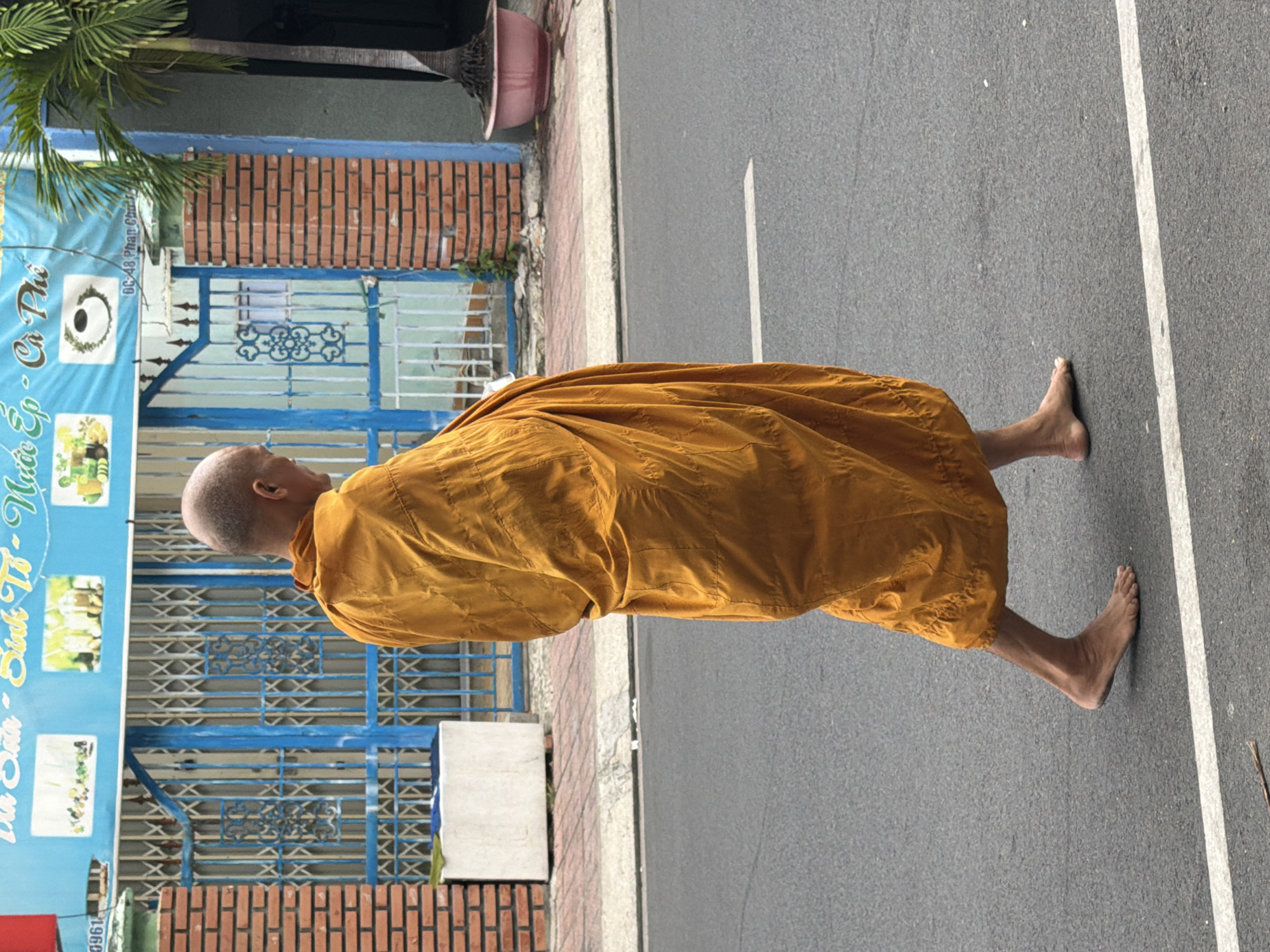 A Buddhist monk in a saffron robe walks barefoot on a paved road in Nha Trang, Vietnam. He is walking from left to right, with his back to the camera. The monk's head is shaved. The road is grey asphalt with a white line painted on it. In the background, there is a building with a blue metal gate and a brick wall. A palm tree is visible to the right of the building. The building appears to be a shop or restaurant, with a sign above the gate that reads "Sinh Tô - Nước Ép - Cà Phê" in Vietnamese, indicating it sells fruit shakes, juices, and coffee. Part of the address "48 Phan Chu Trinh" is also visible on the sign.
