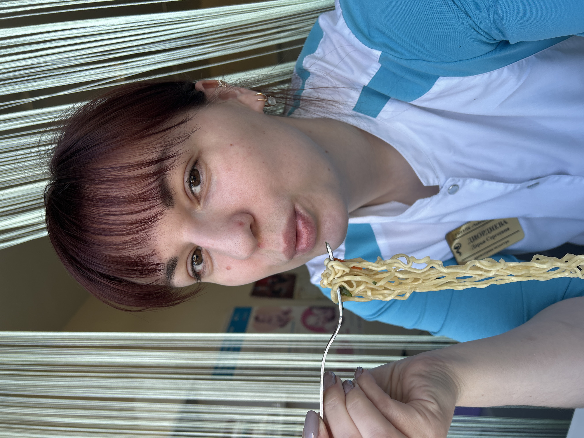 A woman with auburn hair and brown eyes is holding a forkful of noodles near her mouth. She's wearing a white and light blue garment, and a name tag with the Russian text "ДВОРДНЕВА Дарья Сергеевна" (DVORDNEVA Darya Sergeevna) is visible. The location is likely Фоки, Russia, based on the text on the name tag. The background features horizontal blinds or strips. The photo appears to be taken at an angle.