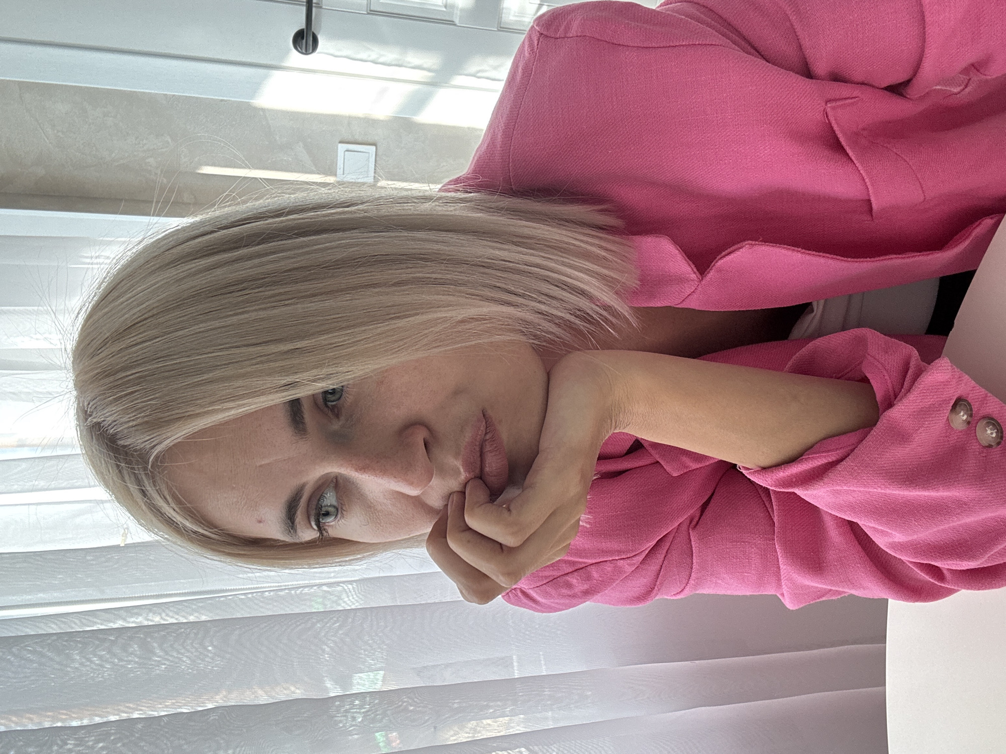 A woman with blonde hair and light blue eyes, wearing a pink blazer, rests her chin on her hand. She is looking to her left, with a contemplative expression. The background consists of sheer white curtains and a wall with a light-colored textured finish, suggesting an indoor setting. There is a door with a black handle on the right side of the frame, and a light switch on the wall. The lighting suggests it is daytime, possibly near a window. The image appears to be a candid portrait, focused on the woman's face and upper body. There is no identifiable information to determine the location within the city of Saratov, Russia.