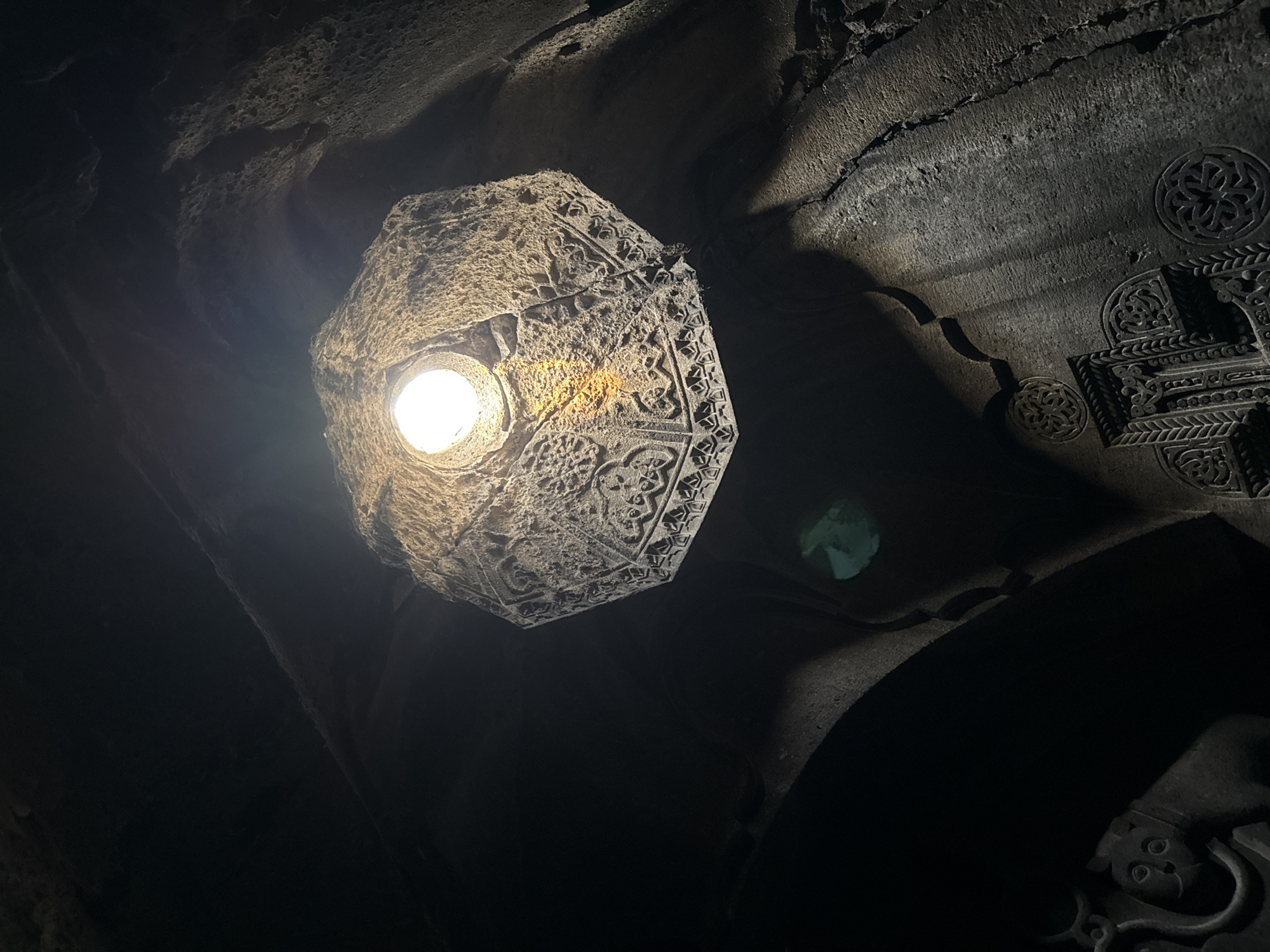 The image shows the interior of a cave or stone structure with a carved ceiling. A single light source illuminates the ornate carvings on the ceiling, which appear to be geometric patterns and possibly stylized figures. The walls are rough and uneven, suggesting a natural cave formation or ancient construction. In the lower left portion of the image, there is a carving of what appears to be a stylized cat or lion. Below that, on the right, are more intricate carvings that resemble crosses and floral designs. The overall impression is one of an ancient, possibly religious, site. This is an interior view of the Geghard Monastery in Goght, Armenia, a medieval monastery recognized by UNESCO.