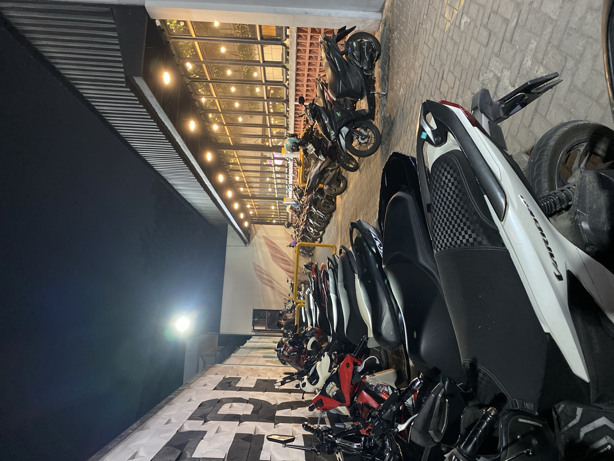 This media file captures a nighttime scene in East Jakarta, Indonesia, featuring a row of parked motorcycles. The lighting suggests it is evening or night, with artificial lights illuminating the area. The motorcycles are parked outdoors, alongside a building with a corrugated metal roof and large glass windows, behind which more lights are visible. The ground is paved with brickwork.
The motorcycles are varied in style and color, predominantly dark, with some red and white accents. They are lined up neatly, indicating a parking area. One motorcycle in the foreground has a white and black body with checkered patterns on the seat. Another has a prominent black front fairing. Some helmets are also visible, resting on or near the bikes. The overall atmosphere is calm and orderly, typical of a parking lot after business hours or in a public space.
There is no visible text in the image, and no people are prominently featured. The focus is on the parked motorcycles and the surrounding architectural and environmental elements. The scene is well-lit by the artificial lights, creating a clear view of the objects.