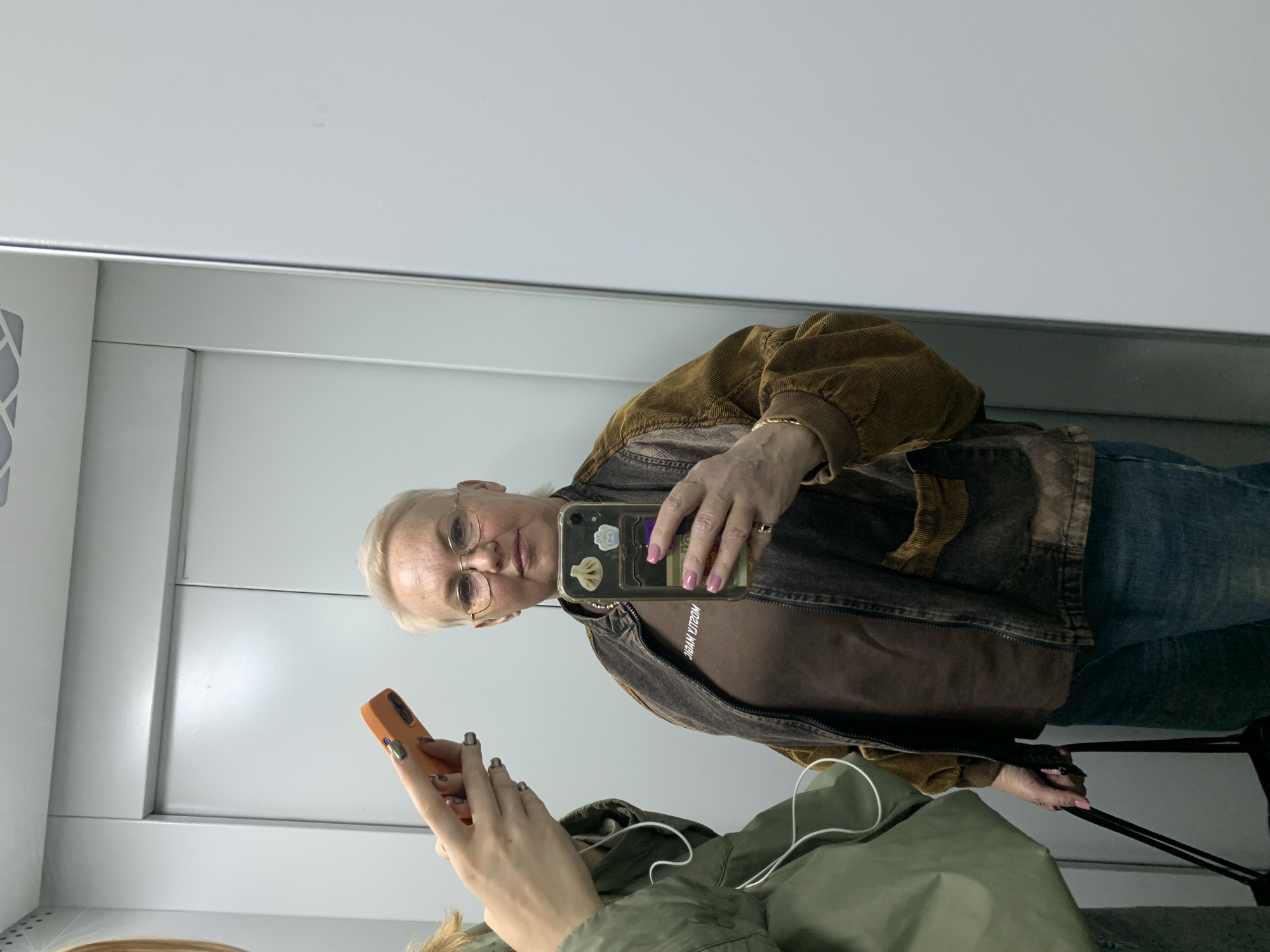 The image shows a middle-aged Caucasian woman with short white hair and glasses. She is wearing a brown jacket over a brown top and jeans. She is holding a smartphone with a clear case that has stickers on the back. She is taking a selfie in a mirror. The mirror reflects a section of a white wall with a seam. Another person is partially visible in the reflection, holding an orange smartphone. The second person is wearing a green jacket. There is a luggage handle visible in the reflection. The image seems to be taken indoors, likely in a commercial space or transportation hub. The location is in the city of Chaikovsky, Russia, based on the image's origin.
