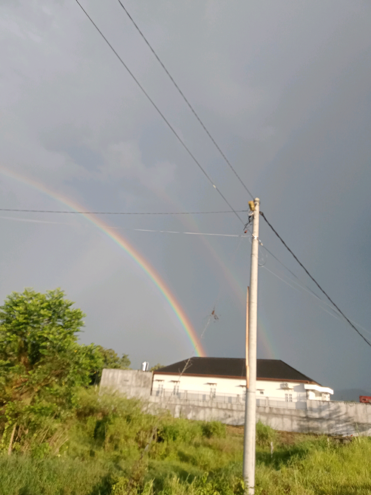 In the city of Hutagaol Peatalum, Indonesia, a double rainbow arcs across a cloudy gray sky, with the brighter primary bow prominently visible. The lower portion of the rainbow extends down to the right, partially obscured by trees. In the foreground, a white utility pole with electrical wires traverses the frame diagonally. To the left of the pole, lush green trees and vegetation dominate the lower half of the image. Behind the vegetation, a white building with a dark roof is visible, bordered by a concrete wall. The overall scene suggests a post-rain or stormy weather condition.