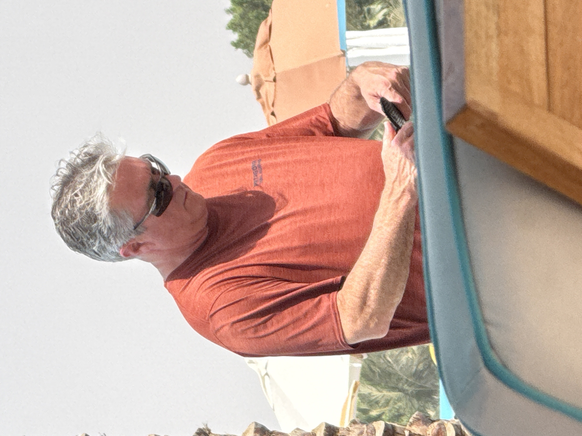 A middle-aged man with graying hair and sunglasses is standing outdoors, looking down at a black object in his hands. He is wearing a reddish-orange t-shirt. The background includes palm trees, a beach umbrella, and what appears to be a swimming pool. The image is taken in Ras Al-Khaimah, United Arab Emirates.