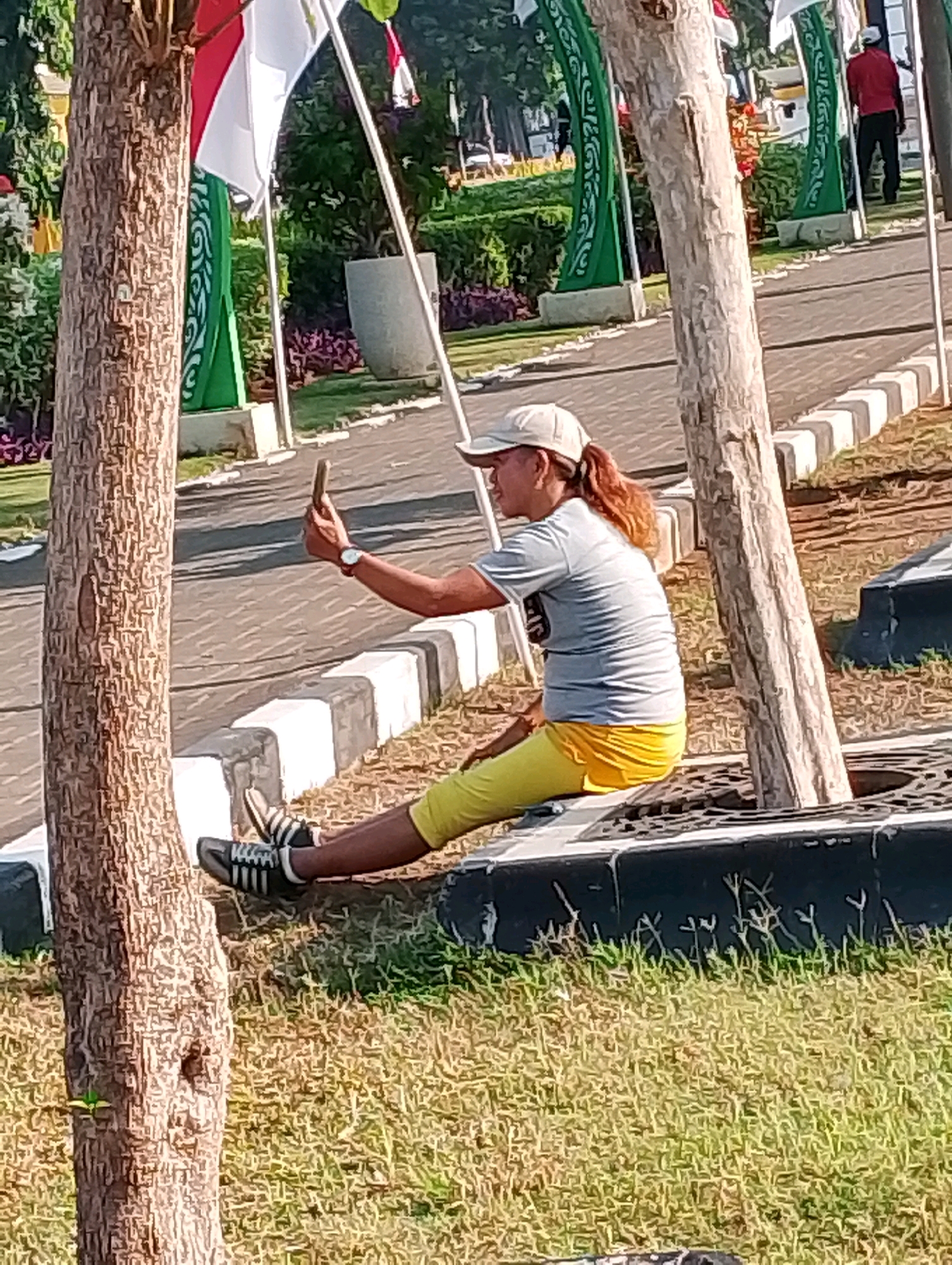 A person is sitting on the edge of a raised circular planter, holding a smartphone in their right hand, seemingly taking a selfie. The individual is wearing a light-colored cap, a gray t-shirt, and yellow pants. Their legs are extended, and they are wearing black and white striped athletic shoes. The person is positioned between two trees. In the background, there are flags, ornamental green banners with white patterns, and a paved pathway with a concrete curb. A person in a red shirt and black pants is visible walking away in the distance. The scene appears to be an outdoor public space, possibly a park or a plaza in the city of Probolinggo, Indonesia. The lighting suggests it is daytime.