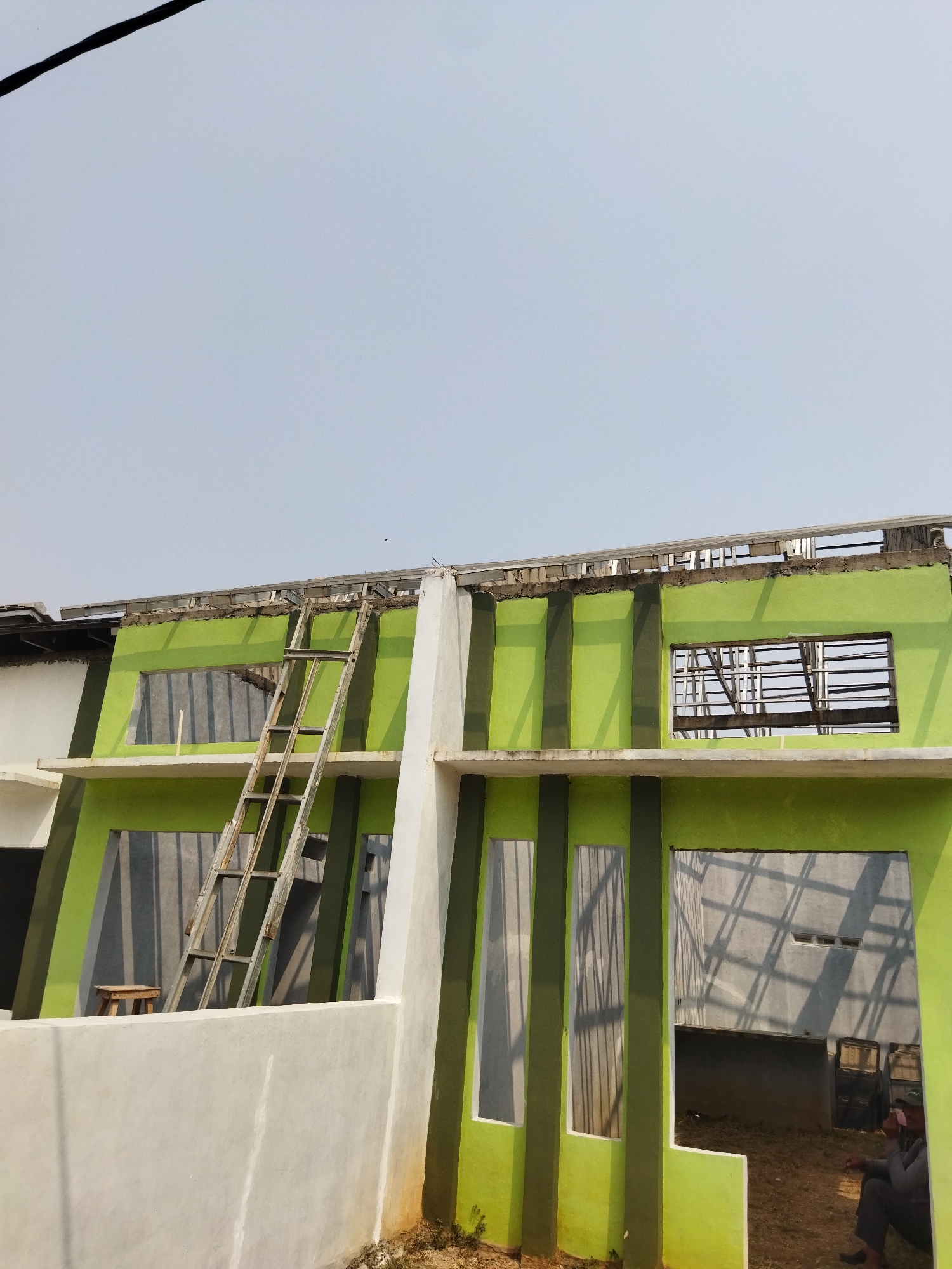 The image depicts the exterior of a two-story building under construction in Suka Sari, Indonesia. The building features vibrant green walls with darker green vertical stripes and white architectural elements. On the left side, a wooden ladder leans against the building, positioned next to a small wooden stool. The windows are open, revealing the skeletal structure within and casting shadows. The rooftop appears unfinished, with exposed metal framing. In the lower right corner, a person is seated on the ground, facing away from the camera, partially obscured by the building's structure. The ground around the building is a mix of dirt and sparse vegetation. The sky is clear and pale blue.