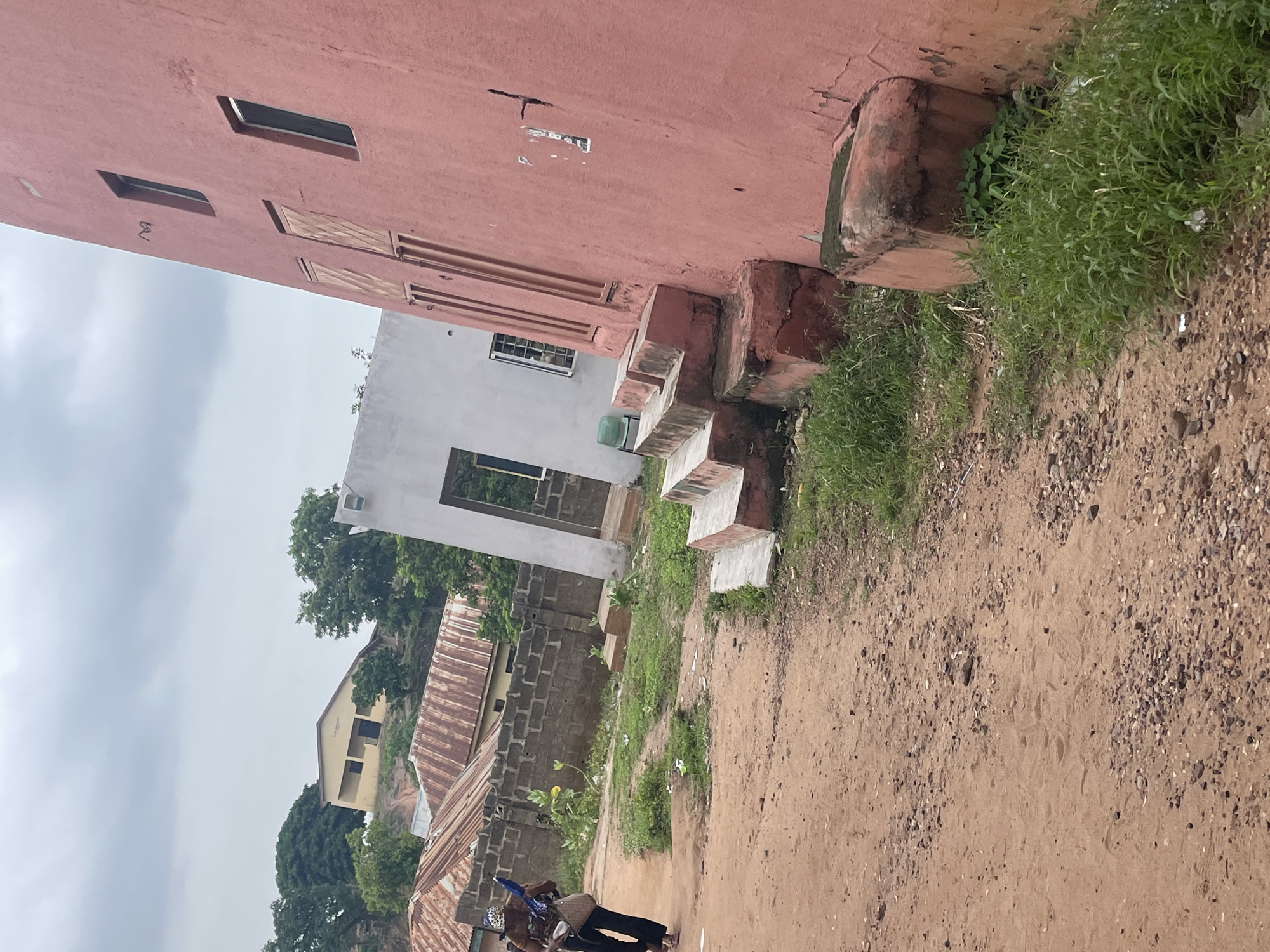 A street scene in Lokoja, Nigeria, depicts a person walking on a dirt road past buildings and vegetation. The road is composed of reddish-brown dirt and small stones, with some footprints visible. To the right, a large, salmon-pink building with multiple windows and what appear to be shuttered doors dominates the foreground. Several concrete steps lead up to an entrance on this building. Adjacent to it, a smaller white building with an open doorway is visible. Behind these structures, a brick wall with corrugated metal roofs of other buildings is present. Further in the background, a yellow building with dark-framed windows is situated on a grassy incline surrounded by trees. Lush green grass and some smaller plants grow along the sides of the road and around the base of the buildings. The sky above is overcast and cloudy.
