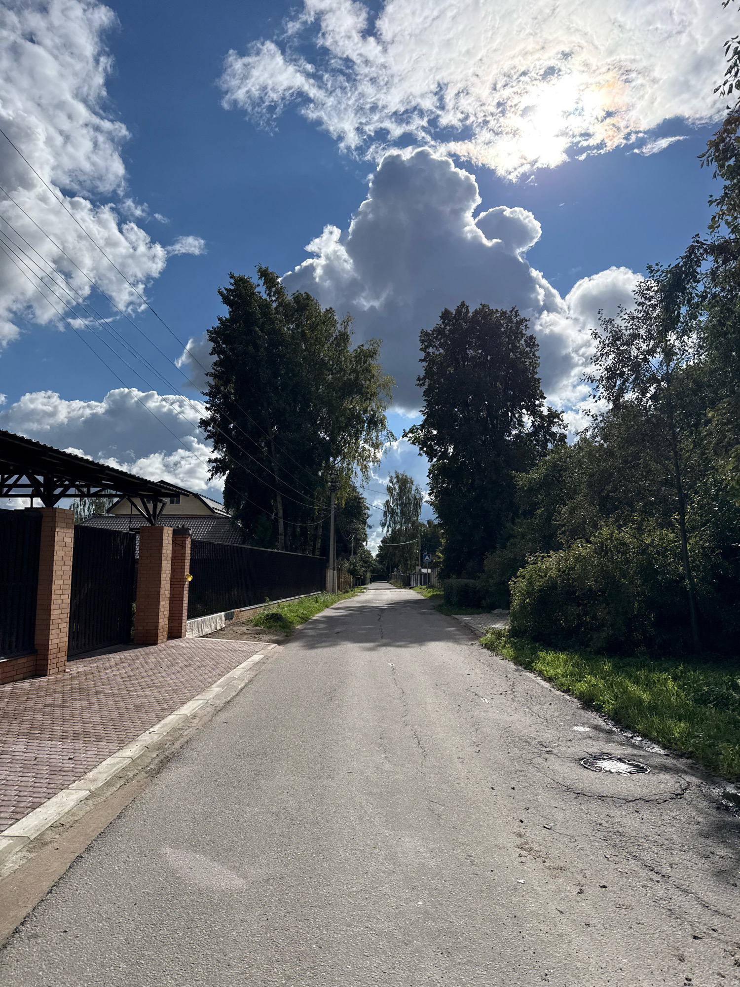 The image depicts a street scene in Vsevolozhsk, Russia. In the foreground, a paved asphalt road runs towards a distant horizon. To the left of the road, a brick and metal fence encloses a property with a covered carport and a house visible behind it. The sidewalk leading up to the fence is paved with red bricks. To the right of the road, a grassy area with trees and bushes lines the street. The sky above is a vibrant blue, dotted with large, dramatic white and gray clouds, with the sun partially obscured by clouds, creating a bright, hazy effect. Electrical wires are strung across the sky on the left side of the image. The overall impression is a sunny, suburban street with some atmospheric cloud formations.