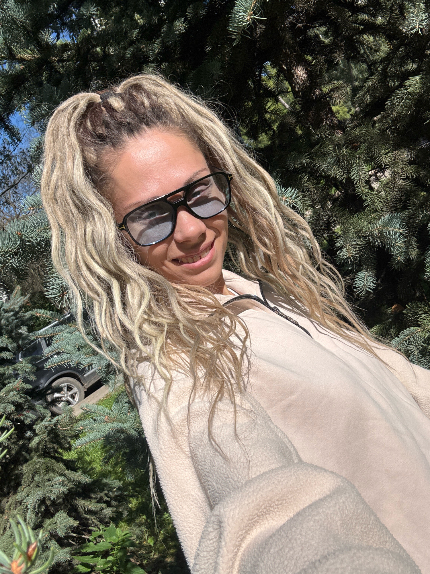 A woman with blonde dreadlocks pulled into a high ponytail, wearing large black-framed sunglasses and a light-colored zip-up jacket, smiles at the camera while standing outdoors. She is partially obscured by dense evergreen trees, with a black car visible in the background among the foliage. The lighting suggests a sunny day, casting shadows and highlights on the scene. The location is not specified, thus it is Unknown, Unknown.