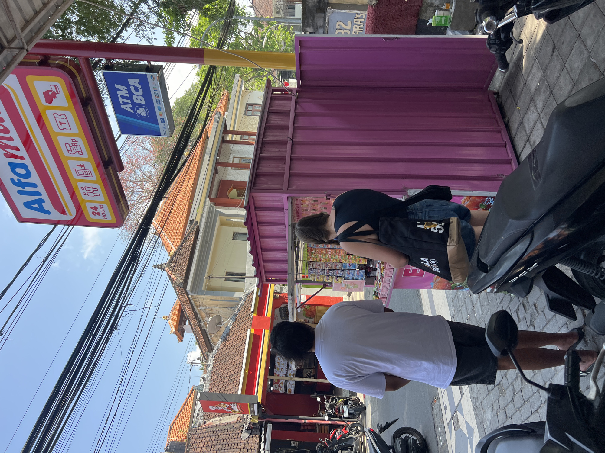 The image depicts a street scene in Kuta, Indonesia, with a focus on a roadside stall and a convenience store. In the foreground, two motorcycles are parked on a paved street. A person with dark hair wearing a dark top and denim shorts stands facing away from the camera, looking at the contents of a pink roadside stall. To their left, a person in a white t-shirt and dark shorts also stands facing away from the camera. The pink stall displays various goods. Behind the stall, a building with a red and yellow awning, possibly a convenience store, is visible. Above this, a bright red and yellow sign for "Alfamart" hangs, along with a smaller blue sign that reads "ATM BCA". Electrical wires are strung across the top of the image against a clear blue sky. The architecture suggests a tropical environment, consistent with Kuta.