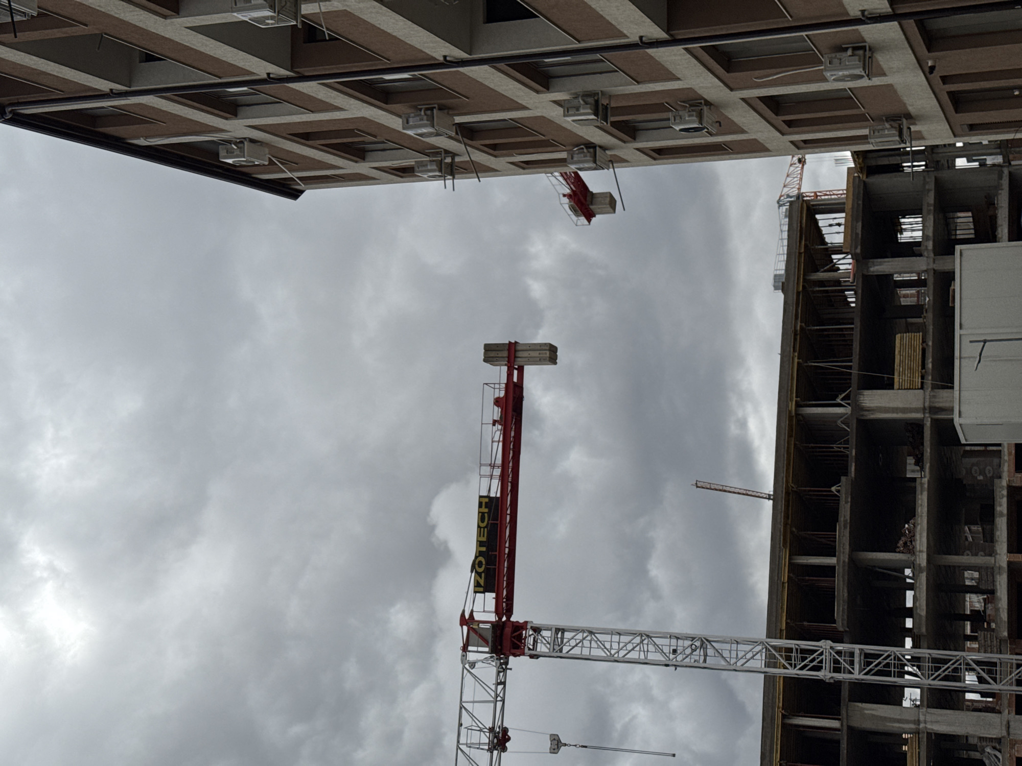 The image shows a view upwards between two buildings in what appears to be a construction zone, against a cloudy sky. On the left is a light-colored building with a grid-like facade. A crane extends over the building on the right, which is an unfinished structure. The name "IZOTECH" is vertically printed on the side of one of the cranes. Given the building style and construction cranes, the location might be a rapidly developing urban area, potentially in Sofia, Bulgaria, given the presence of the IZOTECH brand. No people are visible.