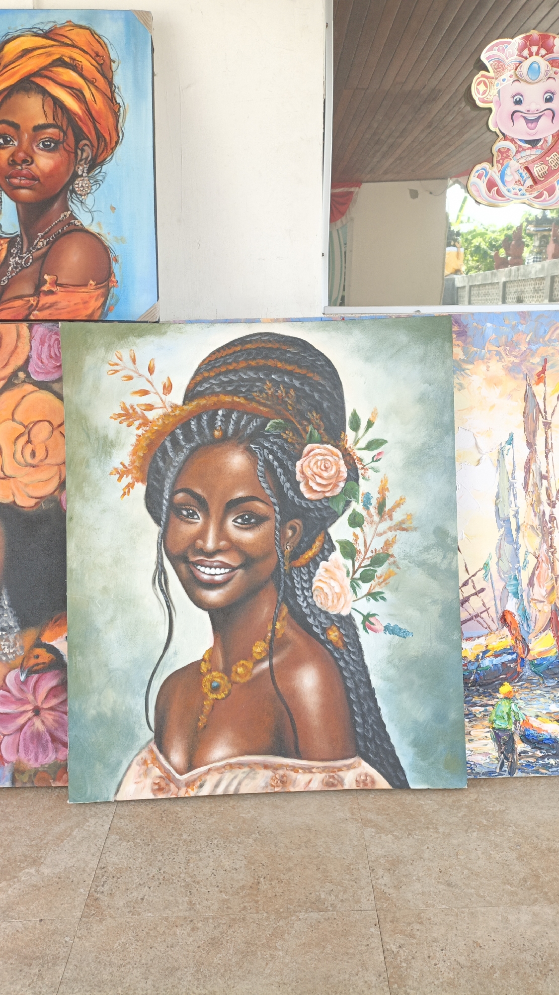 The image displays a collection of paintings displayed outdoors on a tiled floor. The foreground features a large oil painting of a Black woman with her hair styled in braids adorned with flowers. She is wearing a golden necklace and a light-colored garment. To the left of this painting, another artwork depicts a Black woman wearing an orange headwrap and jewelry. To the right, a painting of boats with sails is visible. In the background, a reflection in what appears to be a window or mirror shows a man in a yellow vest and a Chinese deity sticker. The scene is likely located in Sukawati, Indonesia, a known hub for art markets.