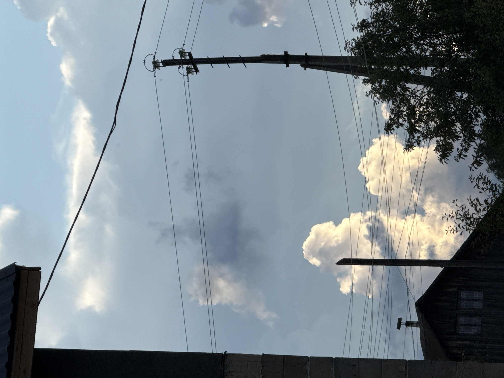 The image depicts a view of the sky with some structures and trees in the foreground, taken from the city of Vetluzhsky, Russia. In the upper left corner, the edge of a dark-colored, corrugated roof supported by wooden beams extends from a dark, vertical structure that appears to be a wall or chimney. A dark electrical wire runs diagonally across the sky from this structure towards the right.
Dominating the sky are large, white, cumulus clouds, illuminated by sunlight from the right, casting shadows on the surrounding sky which is a muted blue with darker gray clouds in the background.
To the right of the frame, a tall, dark electrical pole with multiple crossbars and insulators is visible, supporting several electrical wires that crisscross the sky.
In the lower portion of the image, the dark, wooden roof of a building is visible, with two dimly lit windows and a chimney on top. Adjacent to this building and the electrical pole, dense, dark green foliage of trees fills the right and lower right sections of the frame, obscuring the ground and the base of the structures.