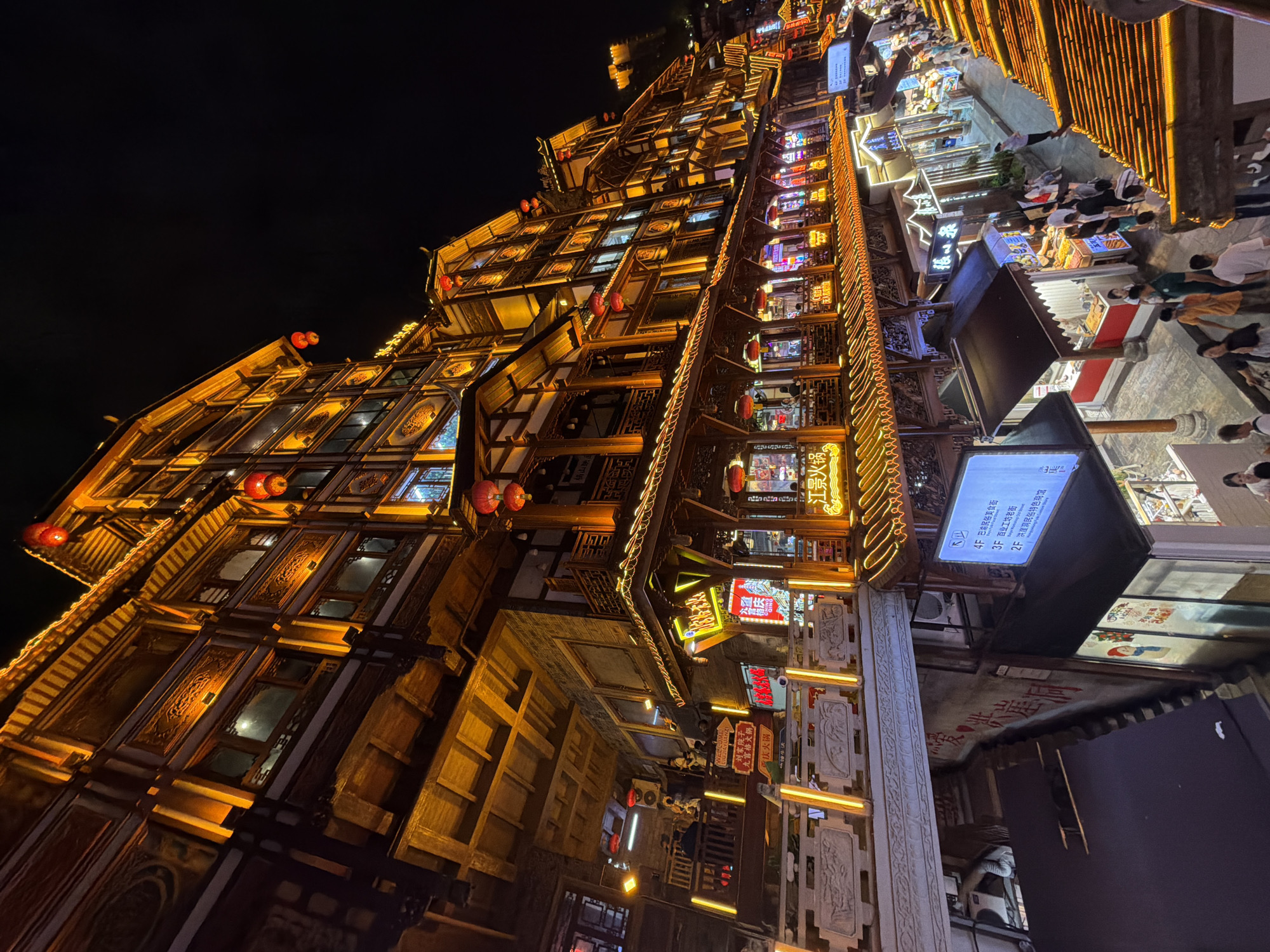 The image captures a vibrant night scene in the Yuzhong District of China, likely a tourist area with traditional architecture. The primary subject is a multi-story building characterized by ornate wooden detailing, tiered roofs, and extensive illumination from warm yellow lights and red lanterns. The building's facade is intricately carved, and its multiple levels are accentuated by balconies and open windows, some of which reveal interiors.
Below the main building, a bustling street scene unfolds. Several smaller stalls and shops are visible, many with illuminated signs. People are walking along the street, browsing the stalls, and gathering in groups. The overall atmosphere is lively and festive, with the architecture suggesting a historical or cultural theme. The lighting is a dominant element, creating a warm and inviting glow that contrasts with the dark night sky. The perspective is from a slightly elevated viewpoint, looking down onto the street and the structures.