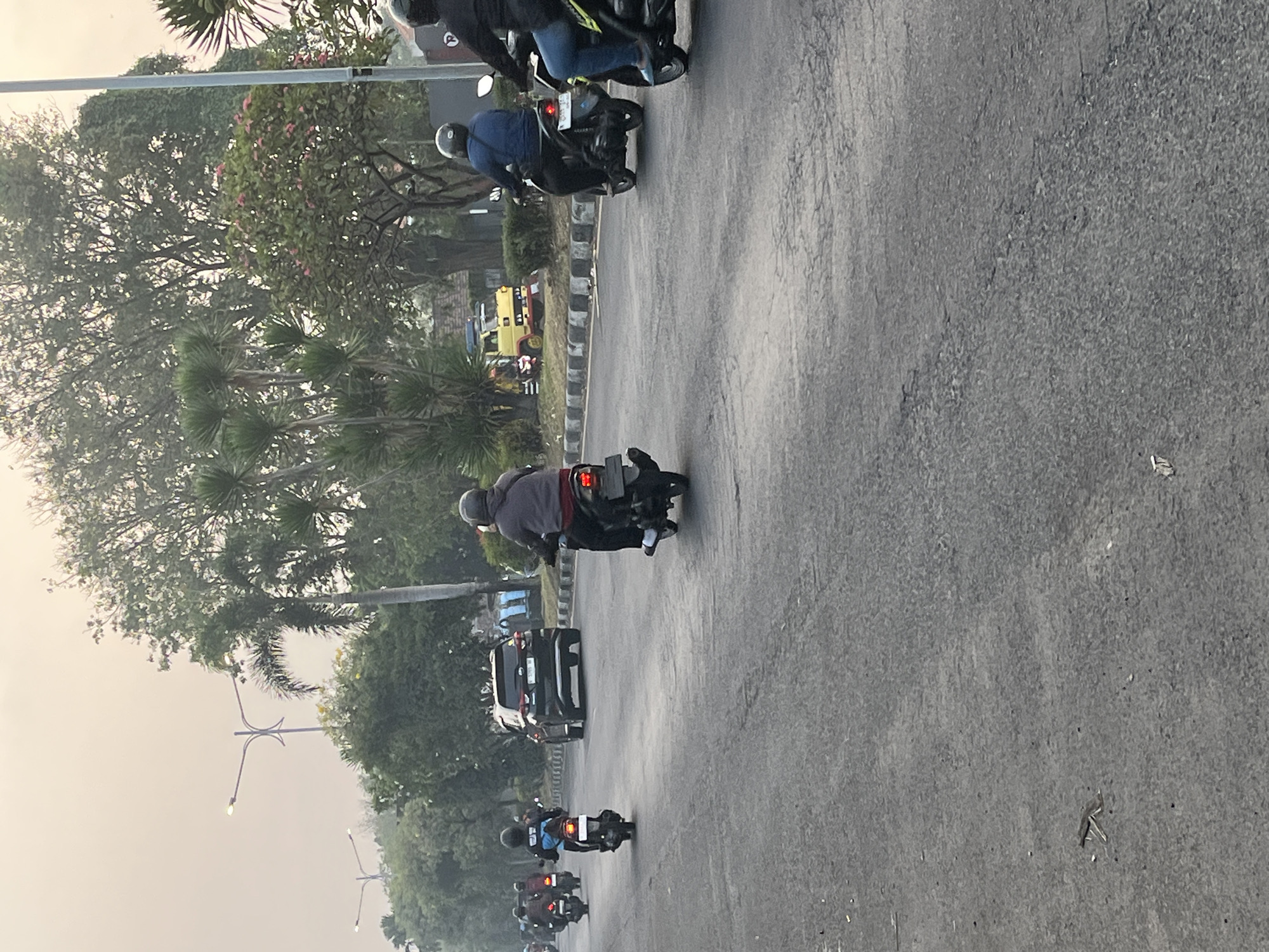 The image depicts a street scene in Kludan, Indonesia, on a hazy day. Multiple motorcycles are visible, with riders wearing helmets, suggesting traffic movement. A black SUV is positioned in the center of the road, its brake lights illuminated. The surroundings include trees, streetlights, and a yellow truck in the distance. The road surface appears to be asphalt.